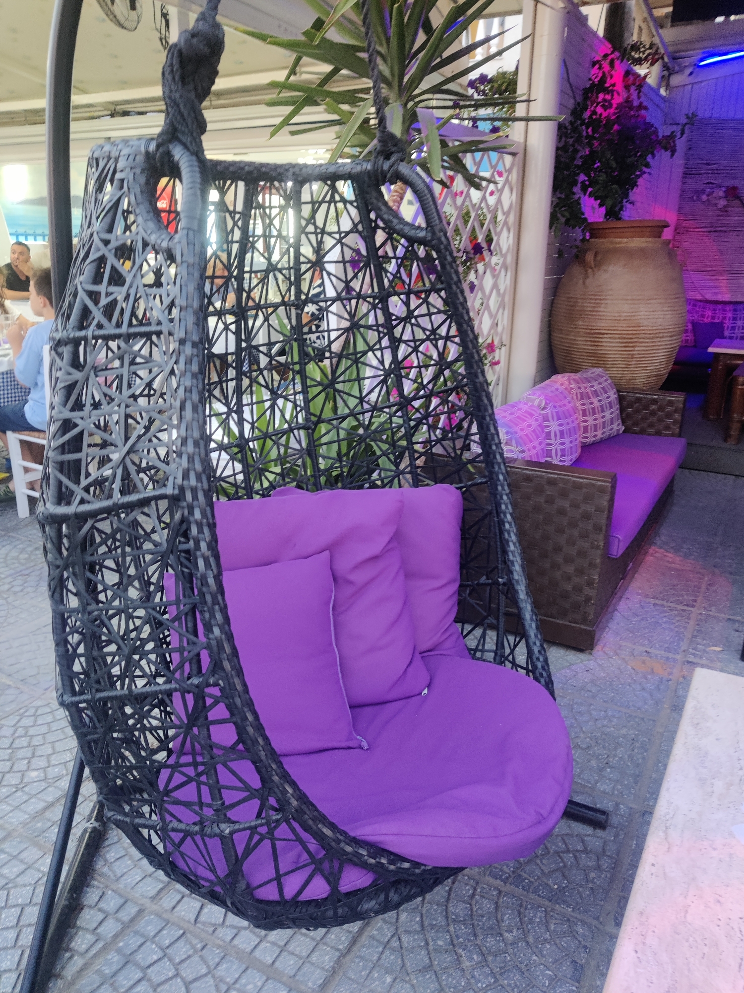 A hanging chair with purple cushions is in the foreground of an outdoor seating area. The chair is made of black wicker and hangs from a metal stand. Behind the chair, a brown wicker sofa with purple cushions is visible, along with a large ceramic pot and a white trellis with pink flowers. The scene appears to be in an outdoor restaurant or cafe in Katerini Municipality, Greece, indicated by the style of furniture and the presence of people dining at a table in the background. The lighting suggests it is daytime.