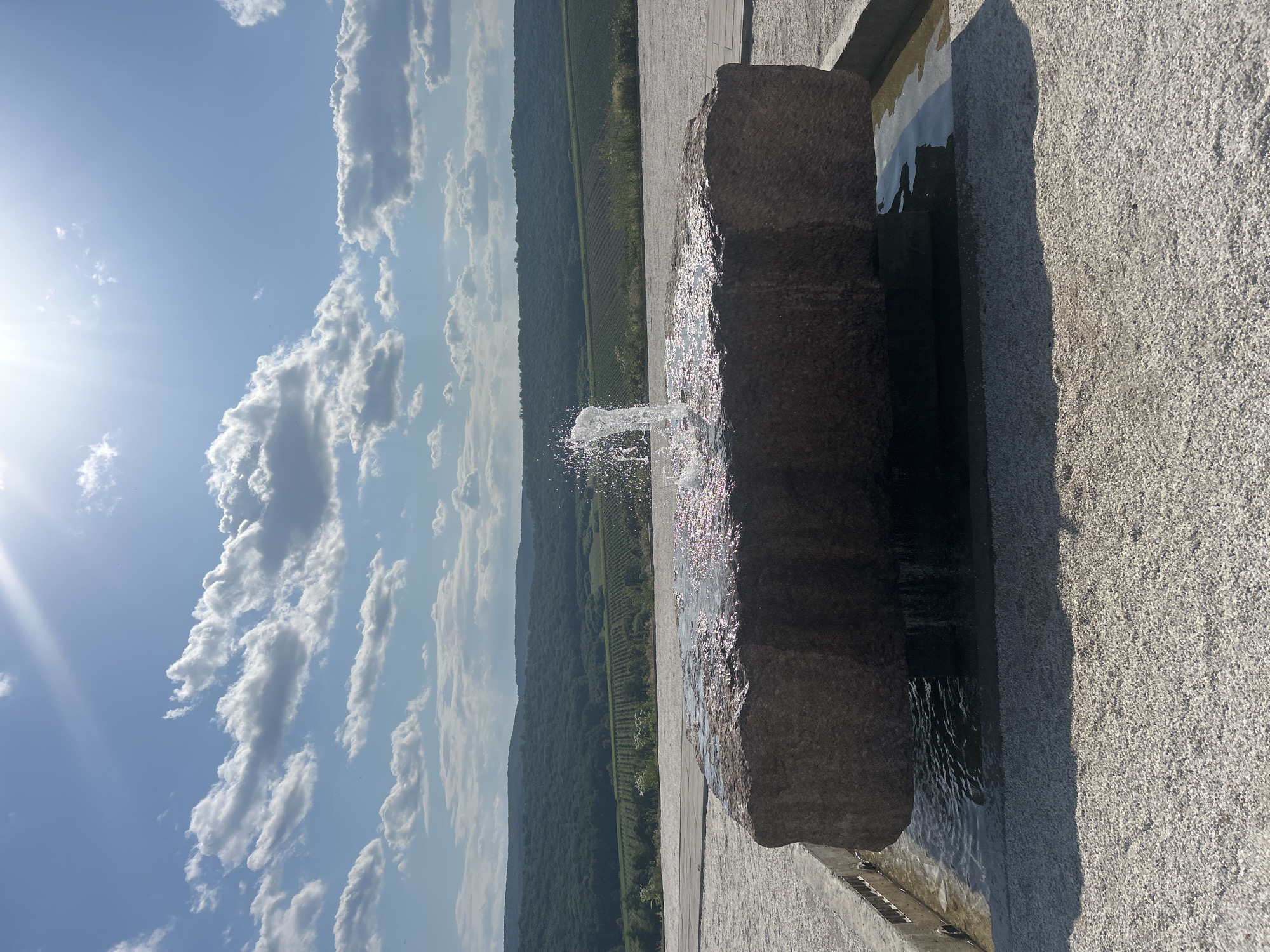 In the city of Ulmu, Moldova, on a bright sunny day with scattered clouds, a contemporary fountain features a large, rough-hewn stone as its centerpiece. Water spouts from the top of the stone, creating a dynamic splash that catches the sunlight. The stone rests in a shallow pool, with water gently overflowing its edges. The immediate surroundings are paved with small, light-colored gravel, creating a minimalist and clean aesthetic. Beyond the fountain area, a paved walkway leads towards a picturesque landscape. Rolling hills covered in dense green trees and vineyards stretch into the distance under a bright blue sky, with the sun creating a lens flare. The scene conveys a sense of tranquility and natural beauty integrated with modern design.