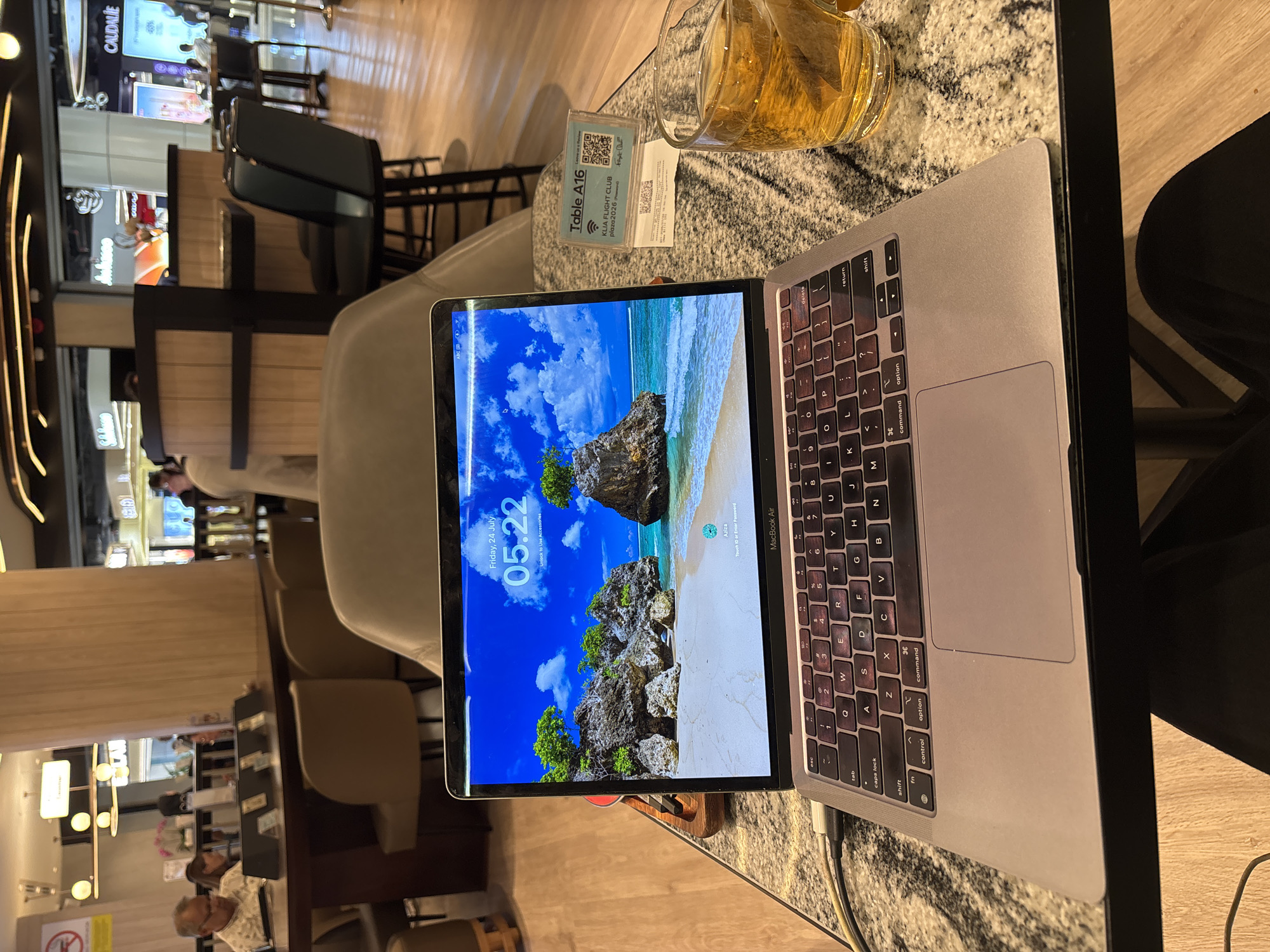 An overhead shot captures a MacBook Air laptop open on a speckled gray countertop table in what appears to be a lounge area. The laptop screen displays a tropical beach scene with a sandy shore, rocky islands covered in lush green trees, and a turquoise ocean under a bright blue sky with scattered clouds. The time on the screen reads "05:22" and the date is "Friday, 24 July".
To the right of the laptop, a clear glass mug filled with golden-brown liquid, likely tea, sits on the table. A tea bag is visible inside the mug. Next to the mug is a small sign with a QR code and text that reads "Table A16", "KLIA FLIGHT CLUB", and "plaza2026 (Password)". A second, smaller slip of paper with similar text and a QR code is beneath the first sign.
In the background, the lounge area features several chairs and tables. A few people are visible, including a man in the left background wearing a patterned shirt, seated at a table. Further back, in the blurred background, are shops and a counter area, suggesting a public space like an airport lounge. The overall lighting is warm and ambient. The scene is set in the city of Sepang, Malaysia, likely within the Kuala Lumpur International Airport (KLIA).