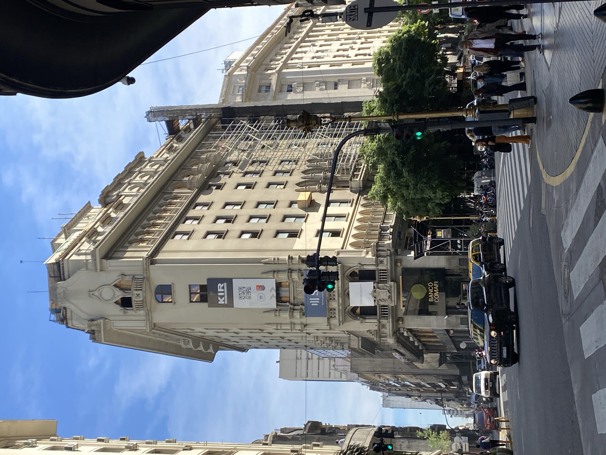 The image captures a street scene in Buenos Aires, Argentina. A tall, ornate building with scaffolding partially covering its upper floors dominates the left side of the frame. A traffic light displays a green signal. Pedestrians are visible on the sidewalk to the right, as well as cars in the street. Street is in front of Banco Comafi.