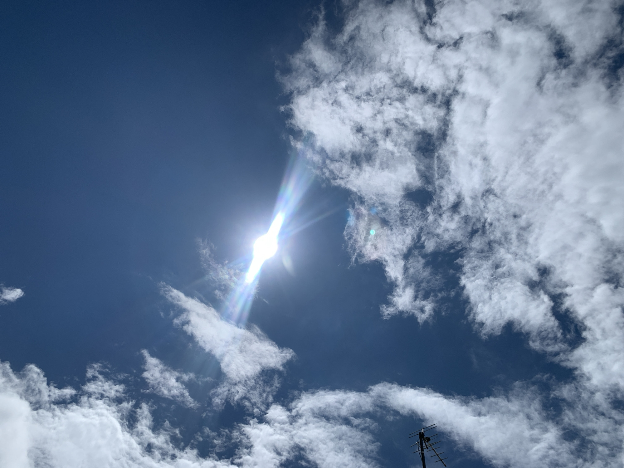A low-angle shot captures a clear, deep blue sky with scattered white clouds. The sun is a bright, lens-flared point in the upper center, emitting strong rays that create a rainbow effect. In the lower left corner, the top of a television antenna is visible, pointing upwards. The scene appears to be an outdoor shot taken during the daytime. The location is specified as Kuru Karama, Nigeria.