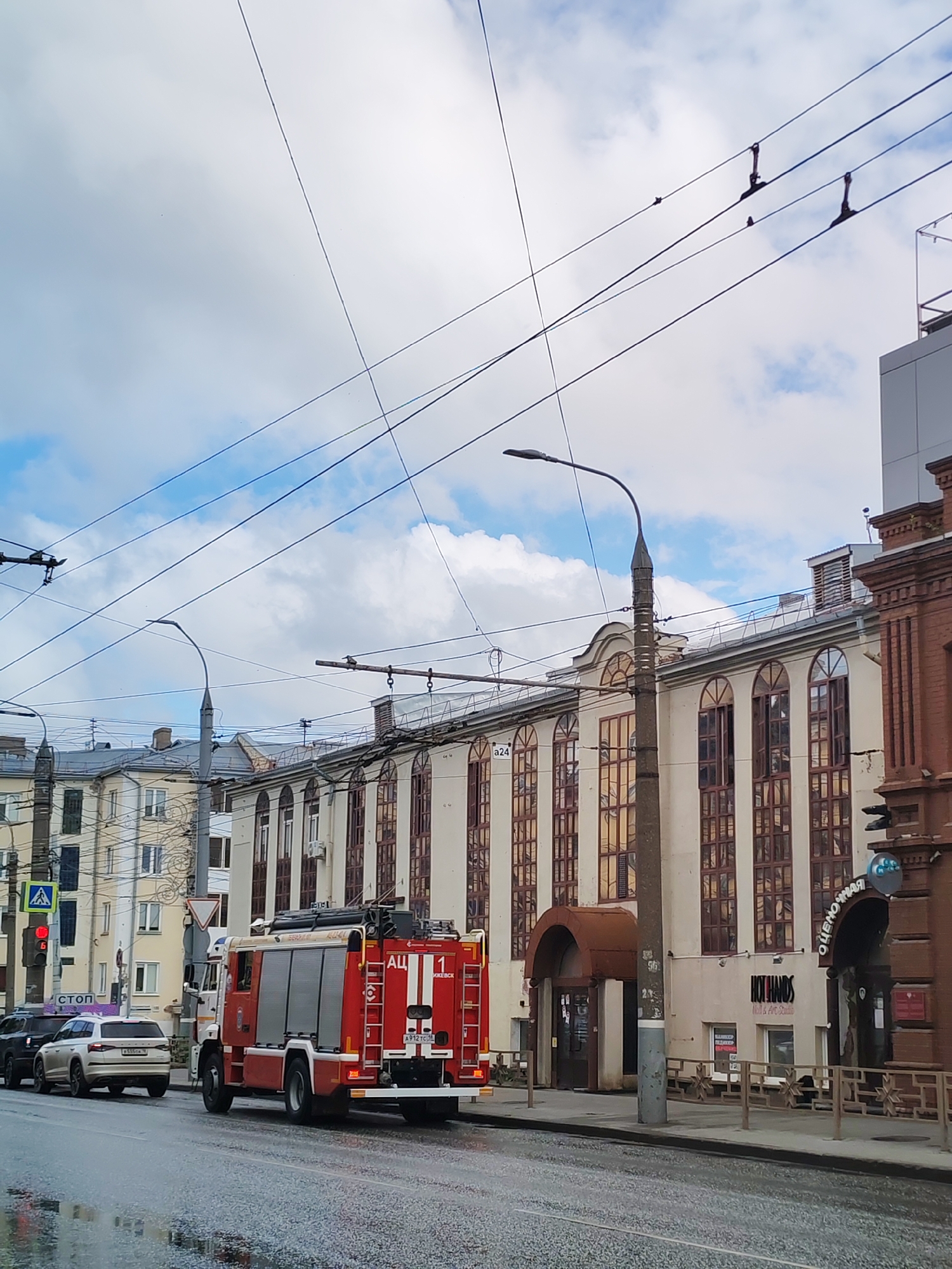 An image shows a street scene in Izhevsk, Russia. A red fire truck is parked on the wet asphalt road, facing left. Behind the fire truck, several cars are stopped at a traffic light. The buildings lining the street have a light-colored facade with large, arched windows. Overhead, a complex network of power lines is visible against a cloudy sky. The scene appears to be during daylight, with overcast conditions contributing to the muted lighting.
