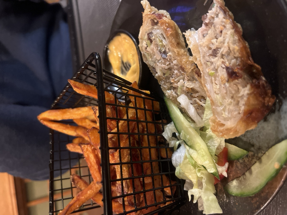 A plate presents two halves of a deep-fried spring roll, revealing a filling of shredded cabbage, meat, and vegetables. Sliced cucumber and lettuce garnish the plate. A small ramekin of orange dipping sauce sits beside the roll. Sweet potato fries, served in a small black metal basket, complete the dish.