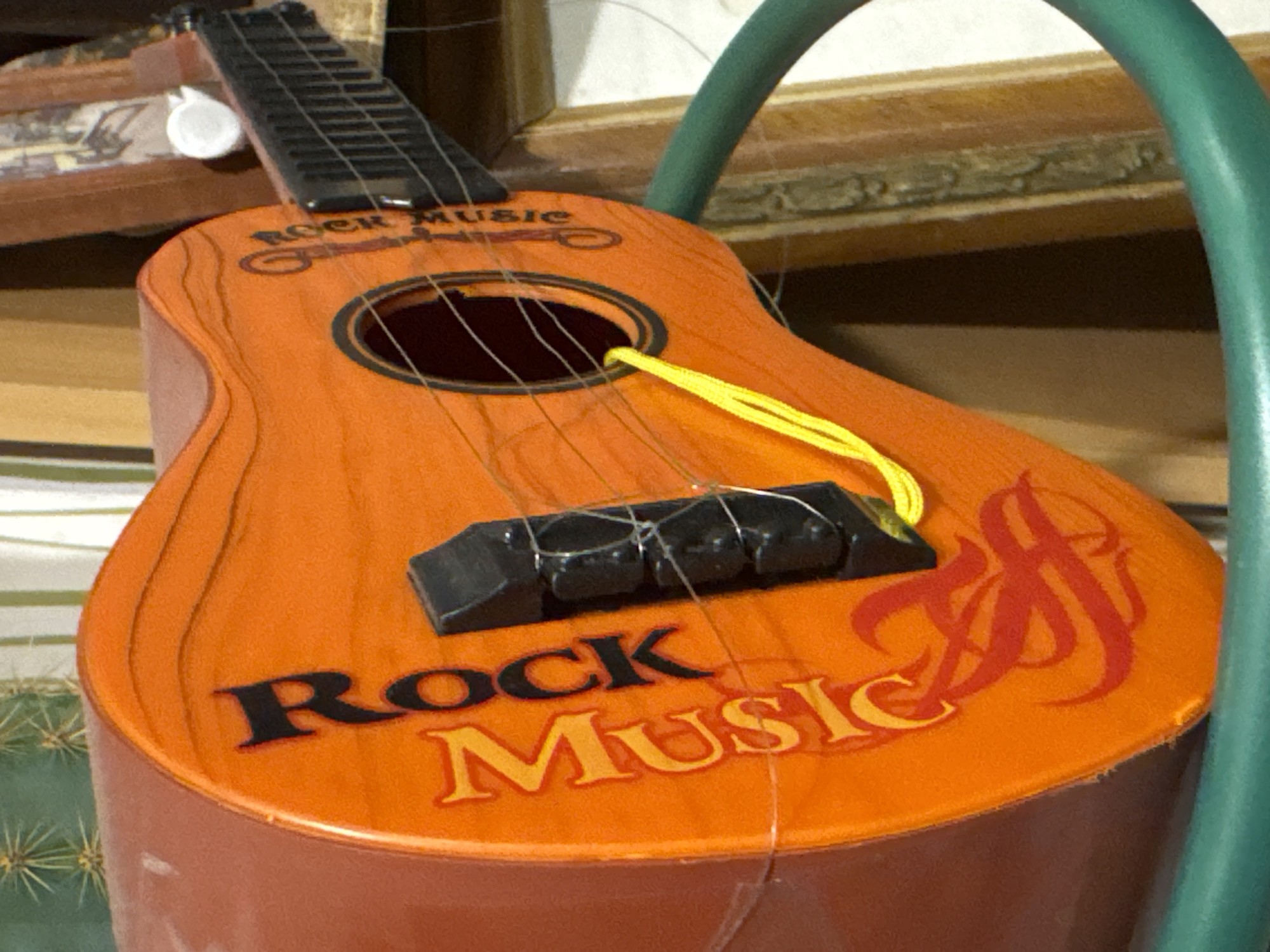The image shows a toy guitar, orange with the words "Rock Music" printed on it. The guitar has a black bridge and a yellow cord running through the soundhole. It appears to be hanging on a green tubular structure. The background includes a wooden shelf with items on it and a plant. It is not possible to determine the location of Ново-Никольское, Russia from the image.
