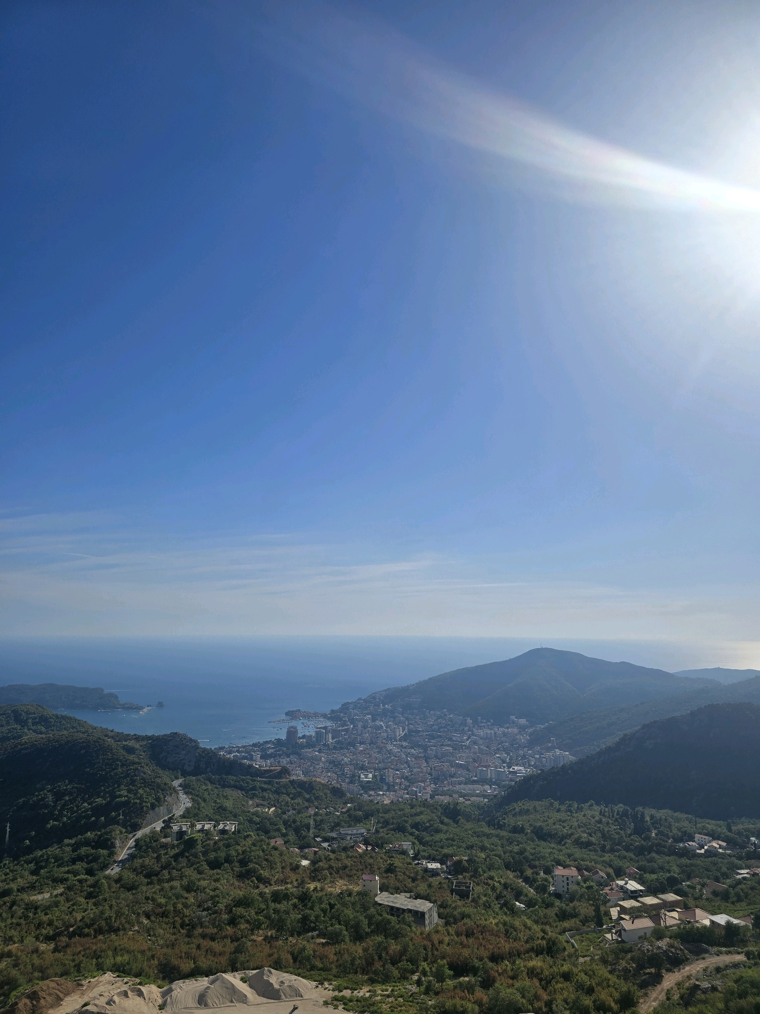 This is a high-angle, wide shot of a coastal city in Lapčići, Montenegro, under a bright, clear blue sky with a sun flare in the upper right quadrant. The foreground shows a dusty, sandy area with mounds of material, suggesting construction or landscaping. Further down the slope, a winding road is visible, with some scattered buildings and dense green vegetation. The mid-ground features a sprawling city with numerous buildings, including taller structures that appear to be hotels or apartments, situated along the coastline. The city is nestled between rolling, tree-covered hills. In the background, the blue expanse of the sea meets the horizon under a hazy sky. The overall scene is a scenic view of a densely populated coastal area surrounded by natural landscape.