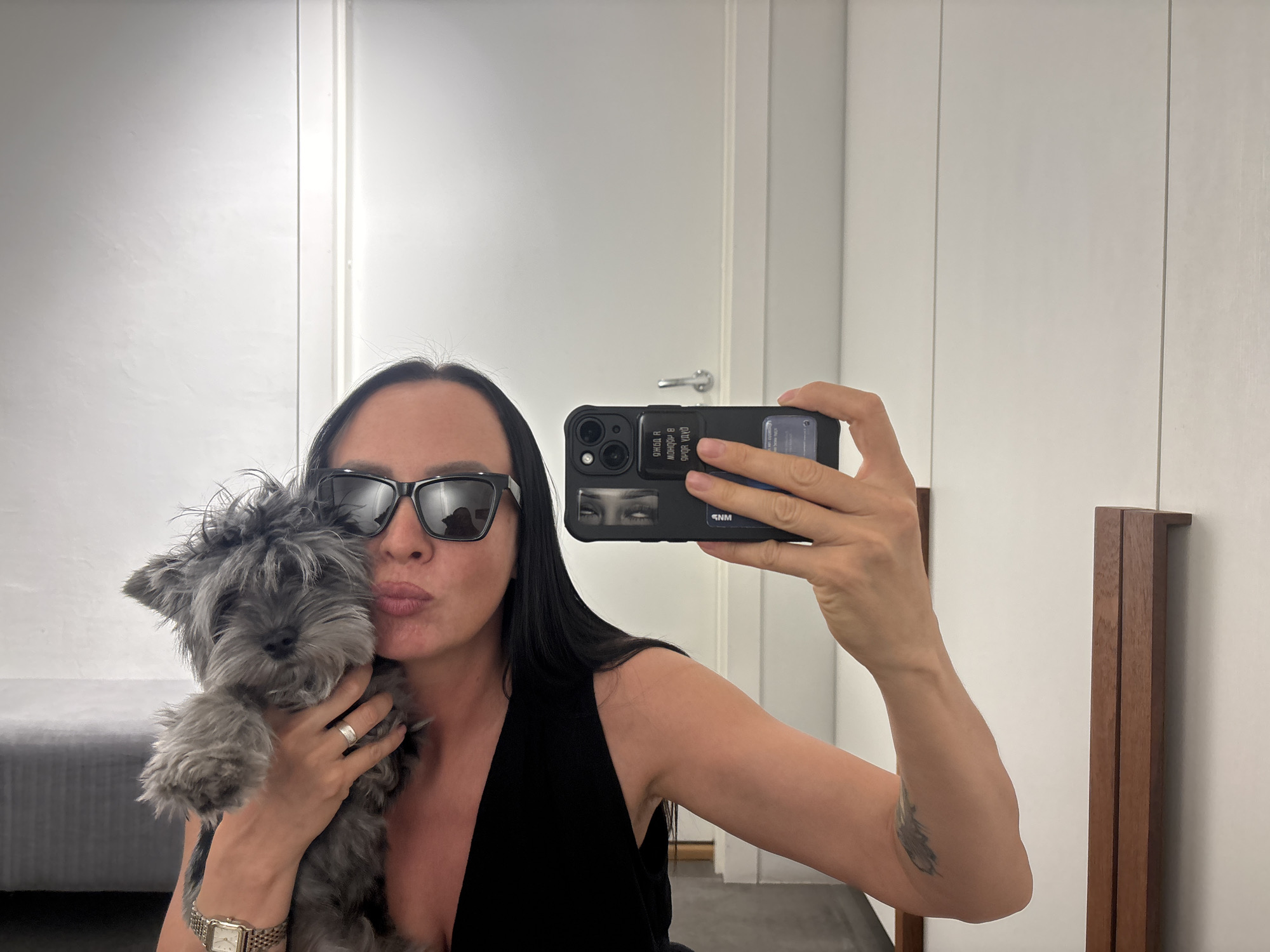 A woman in sunglasses holds a gray dog while taking a selfie in a mirror. The woman has long, dark hair and wears a black sleeveless top. She is kissing the dog's head. The phone case has text printed on it. In the background are white closet doors with silver hardware and a glimpse of a gray bed. The location is Chaikovsky, Russia.