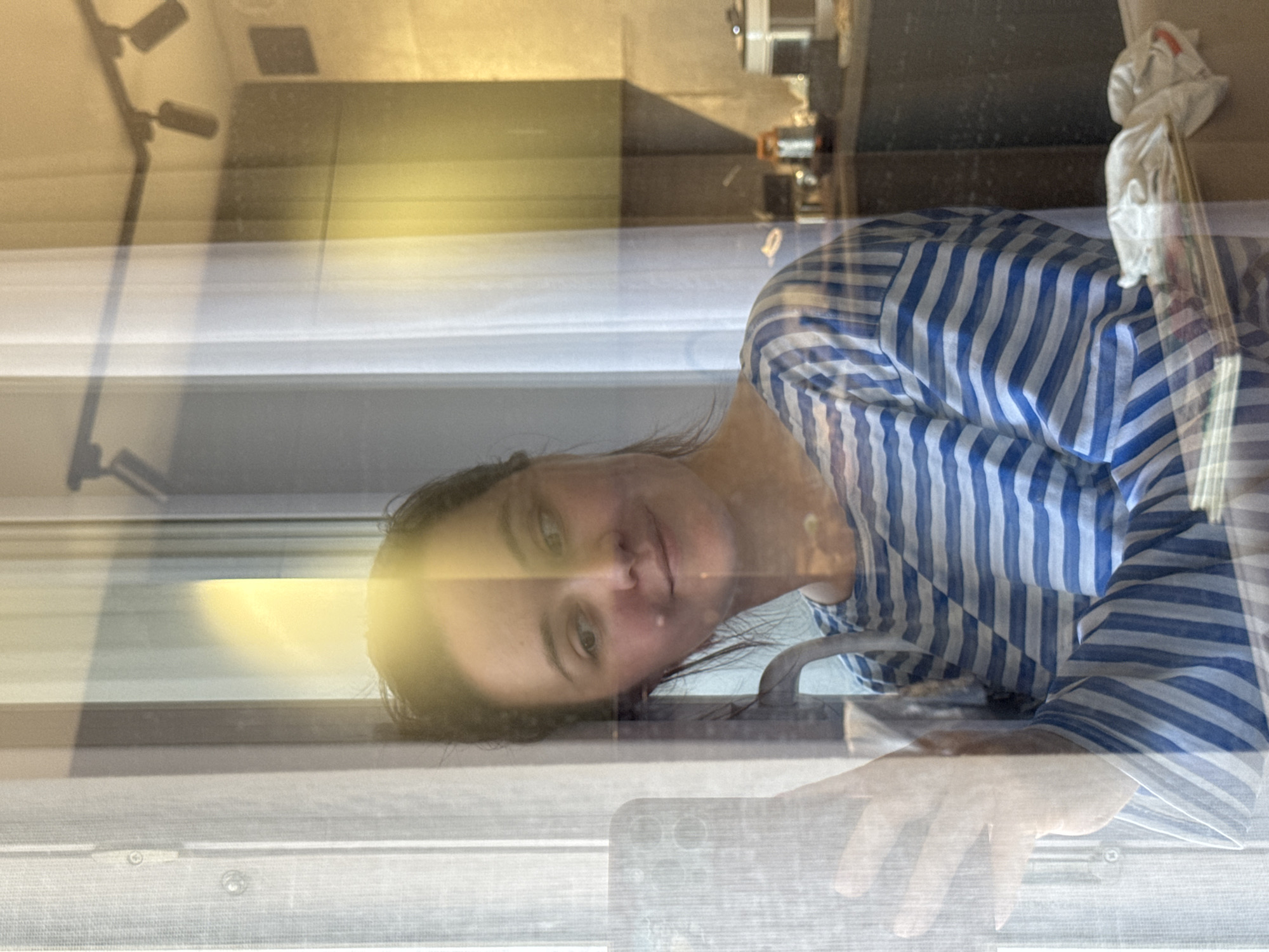 A woman with dark hair and fair skin is seen from the chest up, wearing a blue and white striped shirt. She is looking directly at the camera with a neutral expression. Her hand and a portion of a cell phone are visible in the foreground, seemingly pressed against a window. Reflections on the window reveal glimpses of an interior setting, including what appears to be a kitchen area with cabinets and appliances. Additional reflections show a lamp fixture. The scene is complex due to the window reflections, making it difficult to discern specific details about the setting beyond the kitchen and internal lighting elements. There is no data in the image that reveals the city of Izhevsk, Russia.
