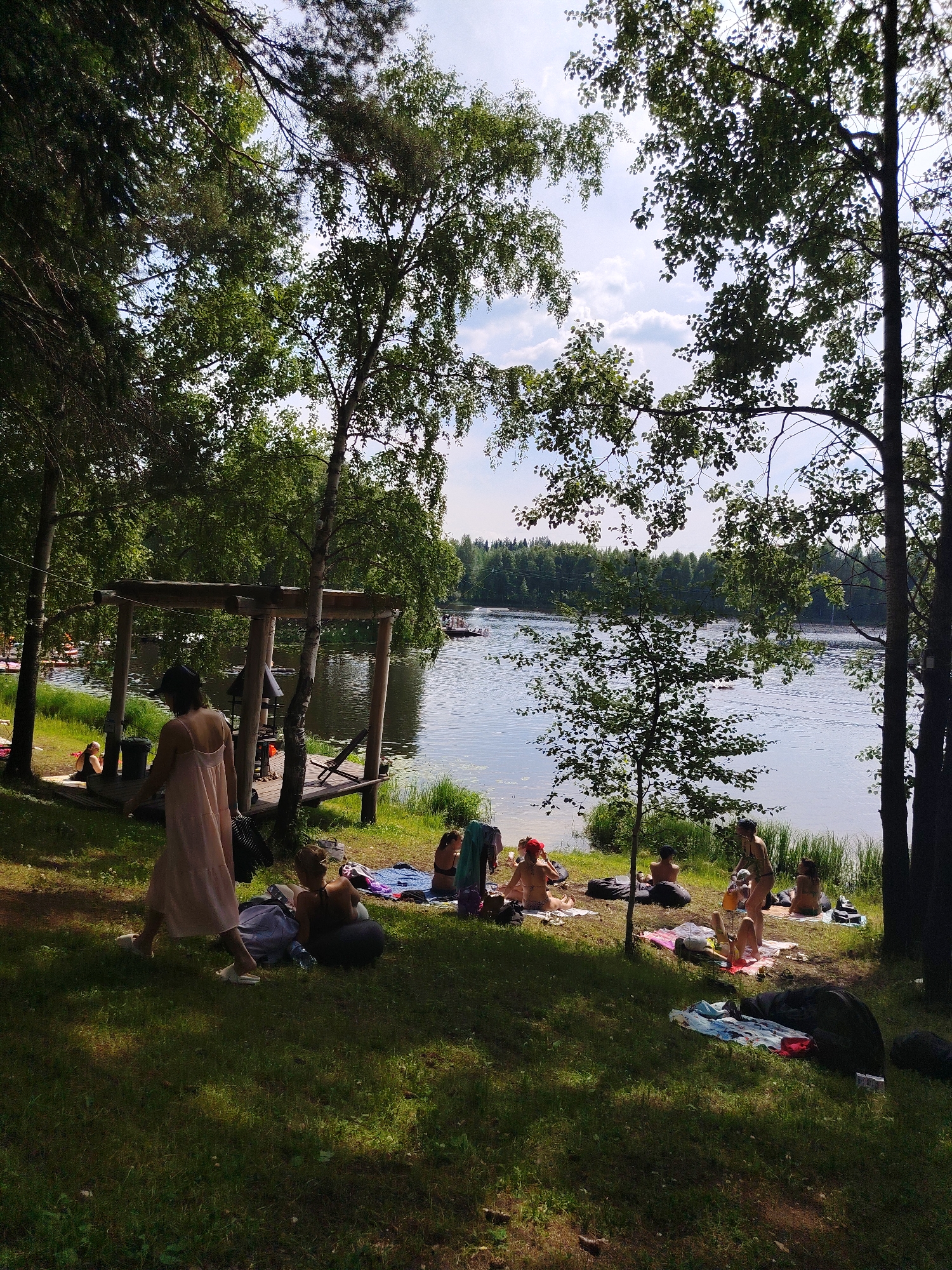 In the city of Vyltydor, Russia, a serene scene unfolds by a lake on a bright, sunny day. The image captures a group of people enjoying leisure time on the grassy shore, shaded by lush green trees.
In the foreground, a woman in a light pink dress walks away from the camera, her white sandals visible on the sun-dappled grass. To her left, another woman is seated on a mat, wearing a black bikini top and shorts. Further to the left, near the lake's edge, a woman with blonde hair sits on a wooden platform, wearing a black swimsuit.
The middle ground reveals more individuals scattered across the lawn, some lounging on blankets and inflatable cushions. A cluster of people are seen near the water, with one woman in a bikini standing and appearing to pour something from a bottle into a glass. Another woman, also in a bikini, is lying on her side on a blanket.
The background is dominated by the tranquil lake, its surface rippling slightly. A wooden structure, possibly a pier or a gazebo, is situated on the left side of the frame, adjacent to the grassy area. In the distance, across the lake, a dense forest line forms the horizon under a partly cloudy sky. There are some boats visible on the lake. The overall atmosphere is one of relaxation and outdoor recreation.