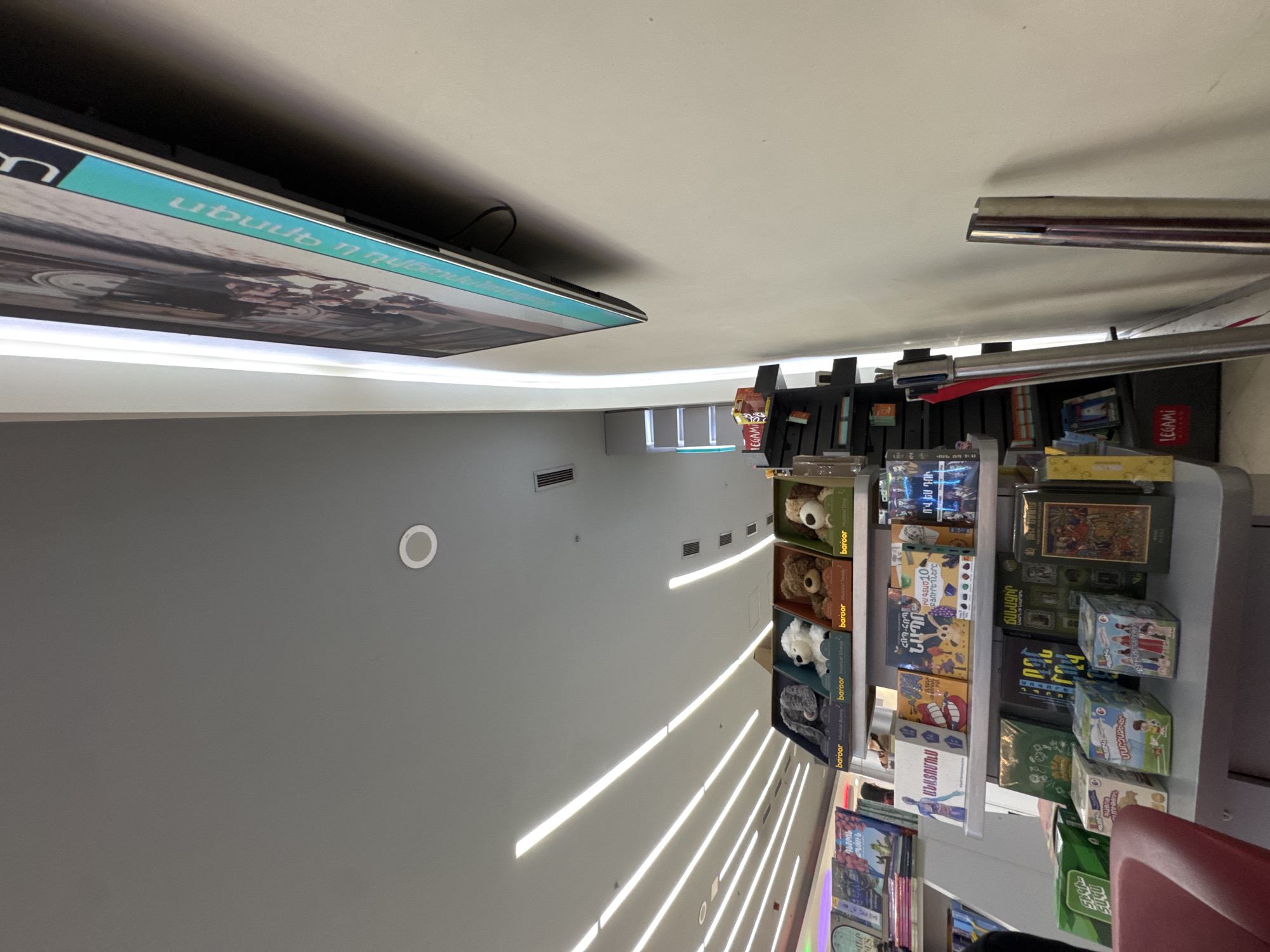 This media file captures an indoor view of a modern commercial space, likely a bookstore or a retail establishment selling toys and games, located in Yerevan, Armenia. The perspective is tilted, showing a bright, high ceiling with numerous parallel LED strip lights. The walls are a neutral grey, also featuring a few of these light strips and a circular recessed light fixture.
A prominent feature is a large, wall-mounted display screen on the upper left, showing imagery and text on a light blue background. Below and to the right, a well-stocked shelving unit displays various products. The top shelves hold plush toys in boxes, including "baroor Musical Dog," "baroor Musical Teddy," and "baroor Musical Sheep." Lower shelves are filled with books and board games.
Visible Armenian text includes titles like "ԴԵՄԲՈՆ" (Dembon), "ԱՆԱՏՈՄՈւՍ" (Anatomus), "ՀՈՊ-ԿՈՊ" (Hop-Kop), "ՆԱՊՈ" (Napo), "ԲՅՈՒՐԵՂՆԵՐԸ" (Crystals), "ՄՈՒԹՅՈՒՆ" (Darkness), "ՄԱՐԶԱՁԵՎԵՐ" (Sports), "ՃԱՆԱՉԻՐ" (Know), "ՔՐԻՍՏՈՆԵՈՒԹ" (Christianity), and "ՎԱՆ ՈՐ ԻՍ" (Van Voris). The text on the large screen's blue banner reads "Սեպտեմբեր 7, ՄԵՆՔ ԵՆՔ ԱՄԵՆԱ..." (September 7, We Are The Most...). The brand name "LEGAMI" (Italian for 'ties', also a brand) is also visible on some product packaging.
The overall scene suggests a clean, well-lit, and organized retail environment, likely aimed at children or families, given the prevalence of games, toys, and educational materials. No people are visible, and the time of day is not discernible due to the indoor setting and artificial lighting.