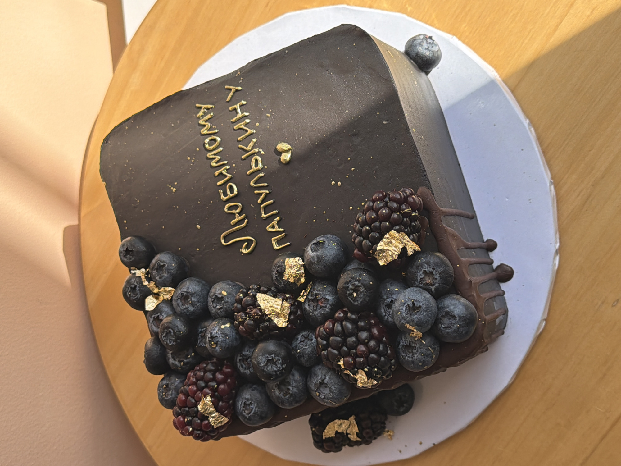 A top-down, eye-level shot shows a square chocolate cake, decorated with blueberries, blackberries, and gold leaf accents, on a white cake board resting on a light-colored wooden surface. The cake has chocolate frosting, and gold lettering is written on the side, oriented vertically. There's a small amount of chocolate dripping down one side. One blueberry is on the top edge of the cake. The location is likely in Tel Aviv, Israel due to the Hebrew lettering on the cake.