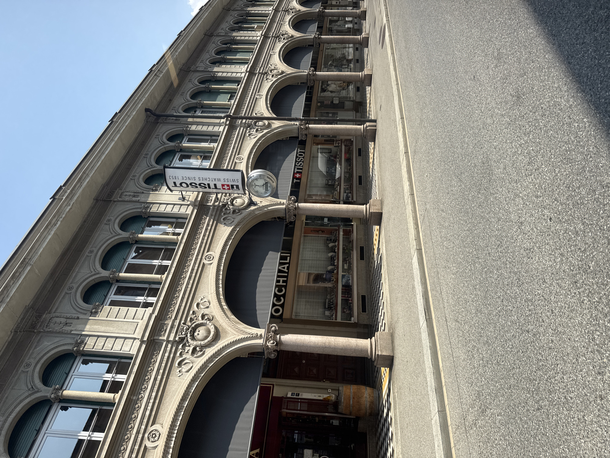 In Lugano, Switzerland, a sunlit street is lined with an ornate, classic building featuring a series of arched arcades. The building's facade is constructed from light-colored stone and adorned with decorative architectural elements, including Corinthian columns and elaborate carvings. Under the arcades, storefronts are visible, one displaying a sign that reads "OCCHIALI" (glasses) and another featuring a Tissot advertisement, with a prominent Tissot clock mounted on the exterior. The ground in front of the shops has a distinctive black and white checkered tiled pattern. A paved road with a grey curb runs along the sidewalk. The sky above is clear and blue.