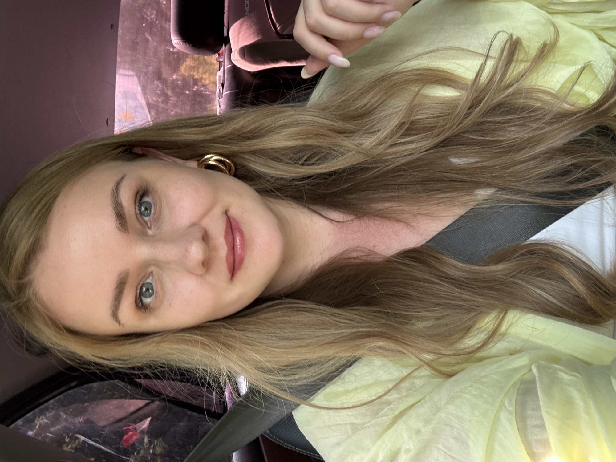 A woman is taking a selfie in a car. She has blonde hair and blue eyes, and is wearing a light yellow top. The background shows the interior of the car and a glimpse of the outside through the window, which suggests a location in a city with trees and buildings.
