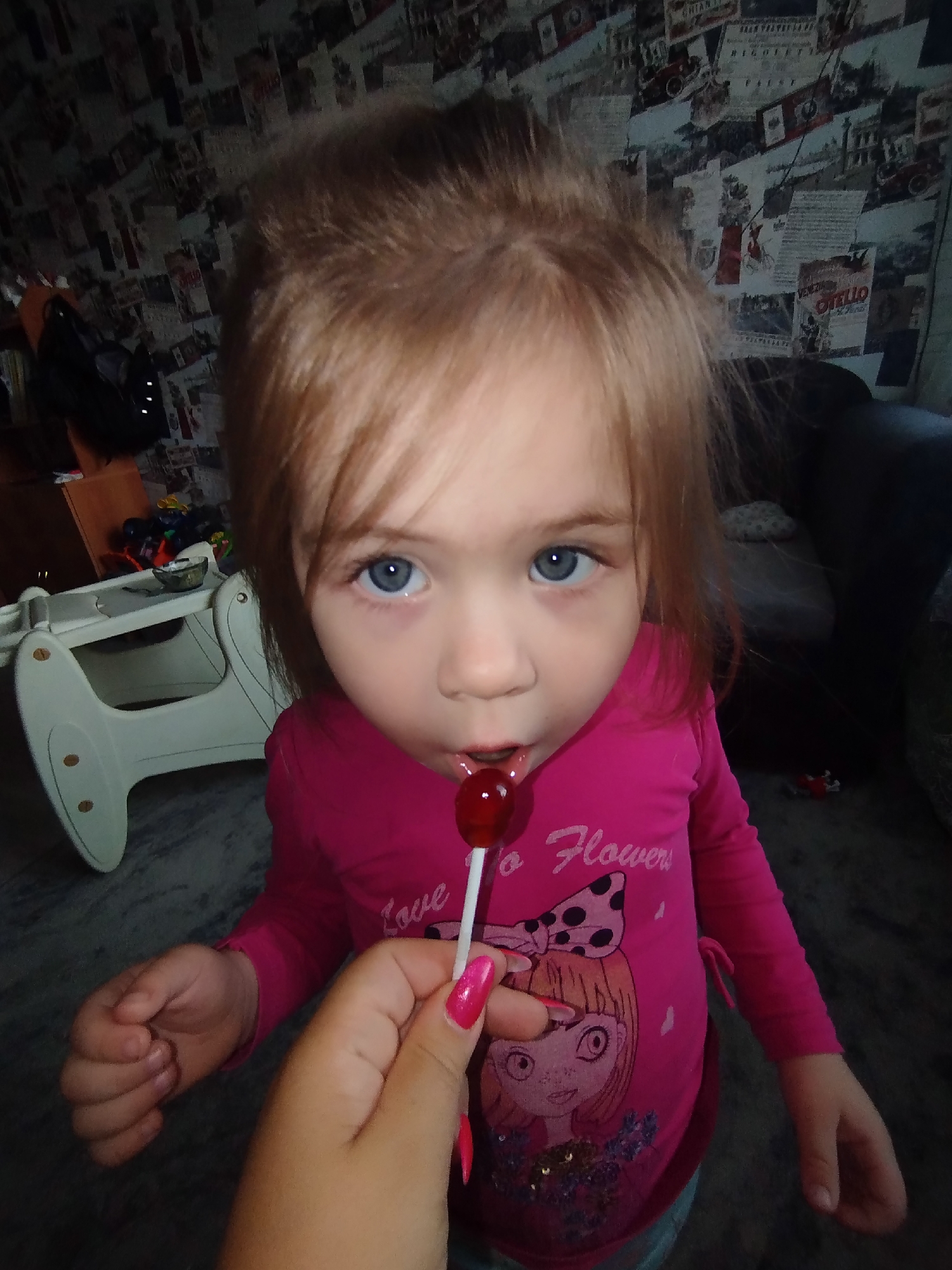 An eye-level indoor shot captures a young child, likely a toddler or preschooler, with light brown hair and prominent blue eyes, looking directly at the camera. An adult's hand, adorned with shiny pink nail polish, holds a red, heart-shaped lollipop on a white stick towards the child's mouth, who is actively engaging with the sweet treat.
The child is wearing a bright pink long-sleeved shirt with the text "Love Flowers" and a graphic illustration of a cartoon girl featuring a large polka-dotted bow. The setting is a room in Chaikovsky, Russia, characterized by a unique wallpaper in the background. This wallpaper is patterned with vintage-style prints, resembling old advertisements, newspaper clippings, or theatrical posters, including visible text such as "VENEZIA," "OTELLO," and "RIGOLETTO."
To the left, a light-colored, possibly white or cream, child's high chair or rocker is partially visible, topped with a small bowl. Dark-colored furniture, possibly an armchair or sofa, occupies the right side of the background. The lighting is diffused, indicating an indoor environment without direct bright light. The child's expression appears focused and curious as they enjoy the lollipop.