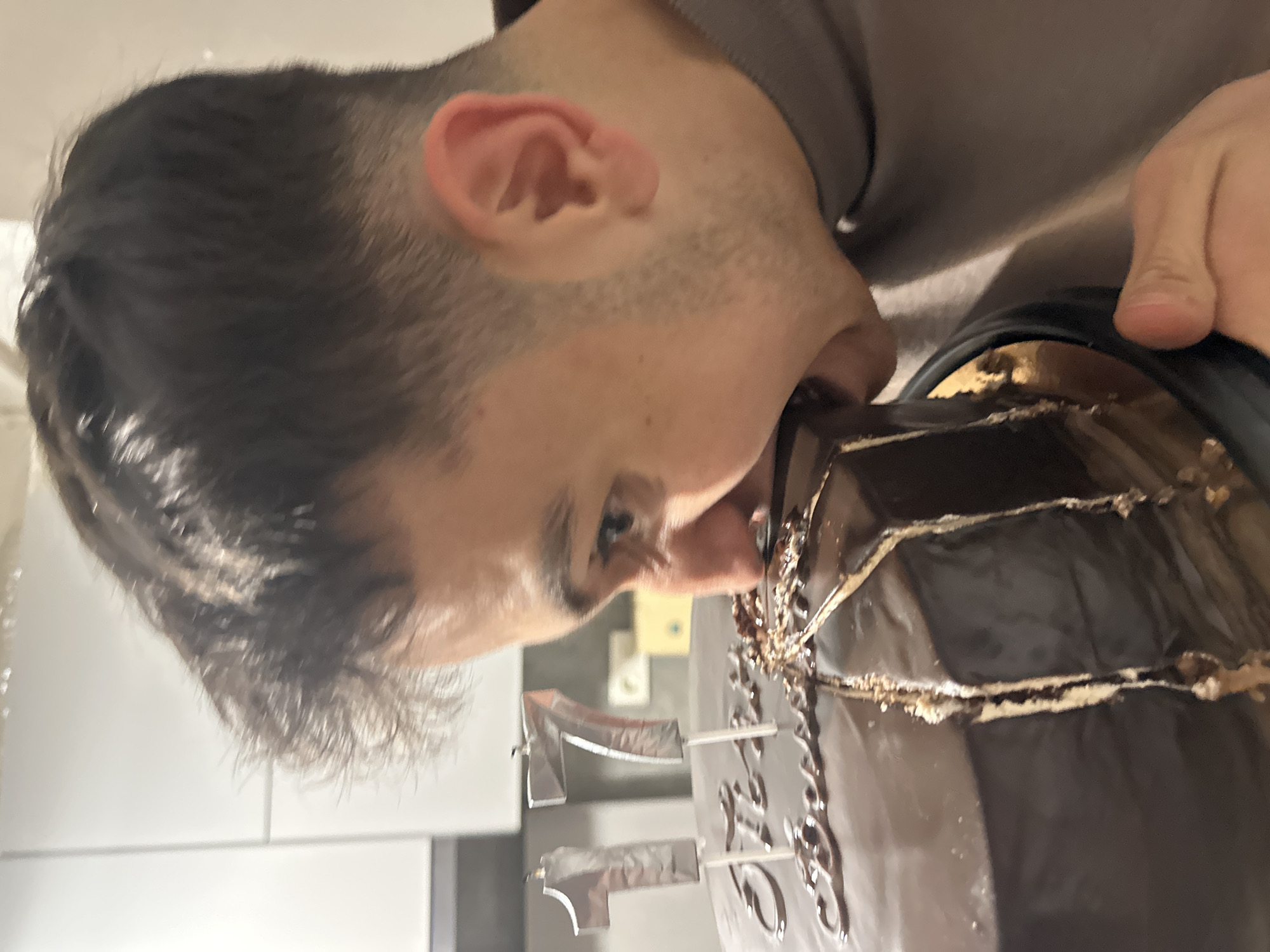 A light-skinned male with short, dark hair is biting into a dark brown, glazed cake with foil number 17 toppers. The background includes light-colored cabinets. The location is Yerevan, Armenia.