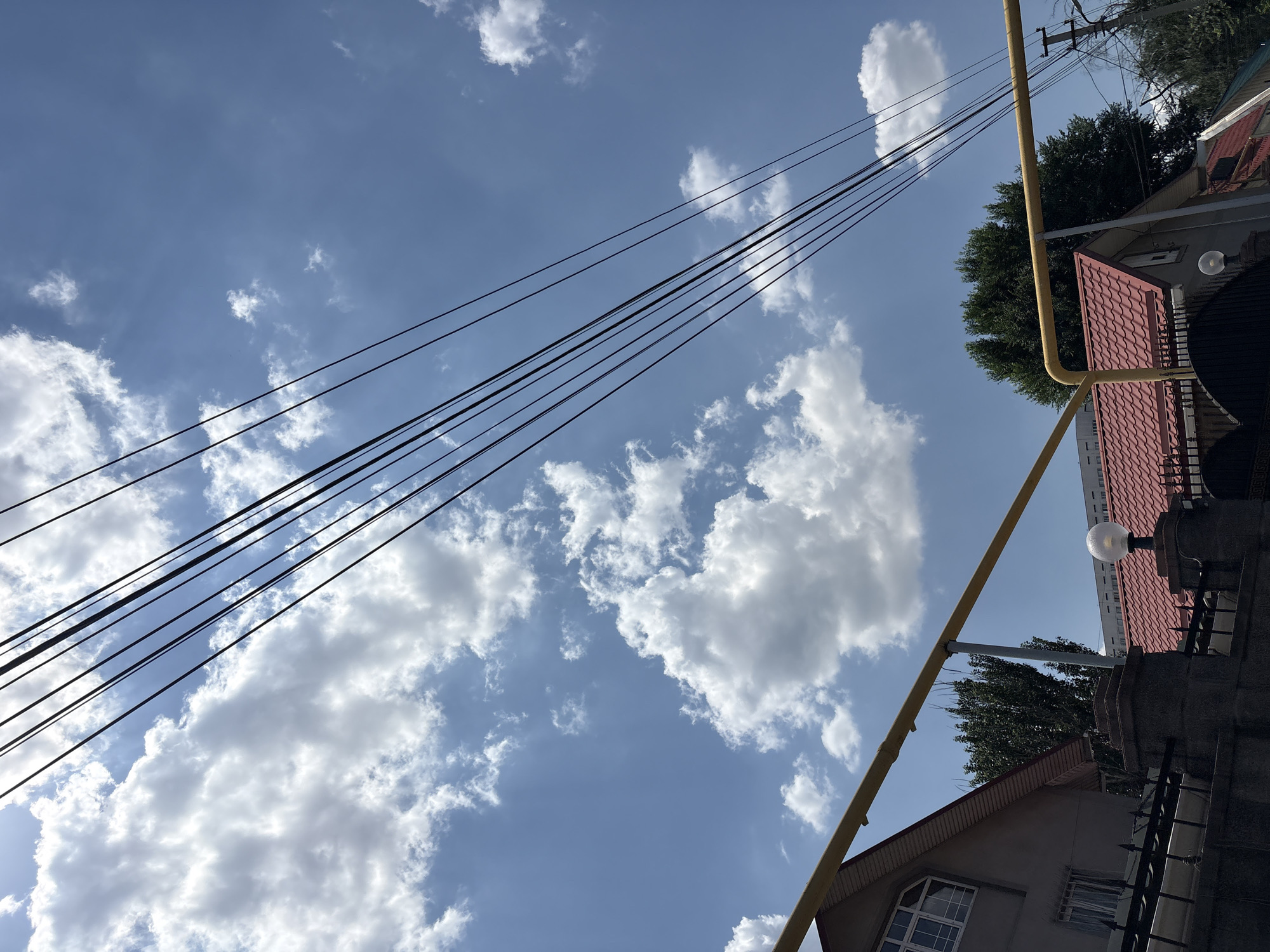 The image captures a bright, sunny day with a blue sky dotted with fluffy white clouds. Several thick black cables stretch diagonally across the upper portion of the frame, obscuring parts of the sky. In the lower half of the image, the rooftops of several buildings are visible, characterized by red tiled roofs and some grey, stucco-like walls. A prominent bright yellow pipe runs horizontally across the middle of the frame, supported by metal poles. To the left, a portion of a grey house with a gabled roof and a window is visible behind a black metal fence. To the right, a lamppost with a spherical light fixture stands atop a stone pillar, also behind a fence. Trees with dark green foliage are present in the background, partially obscuring other buildings. The overall scene suggests a residential area in a city. The architectural style of the buildings, particularly the red tile roofs, and the presence of mature trees are consistent with neighborhoods found in Almaty, Kazakhstan, especially in the older or more established districts.