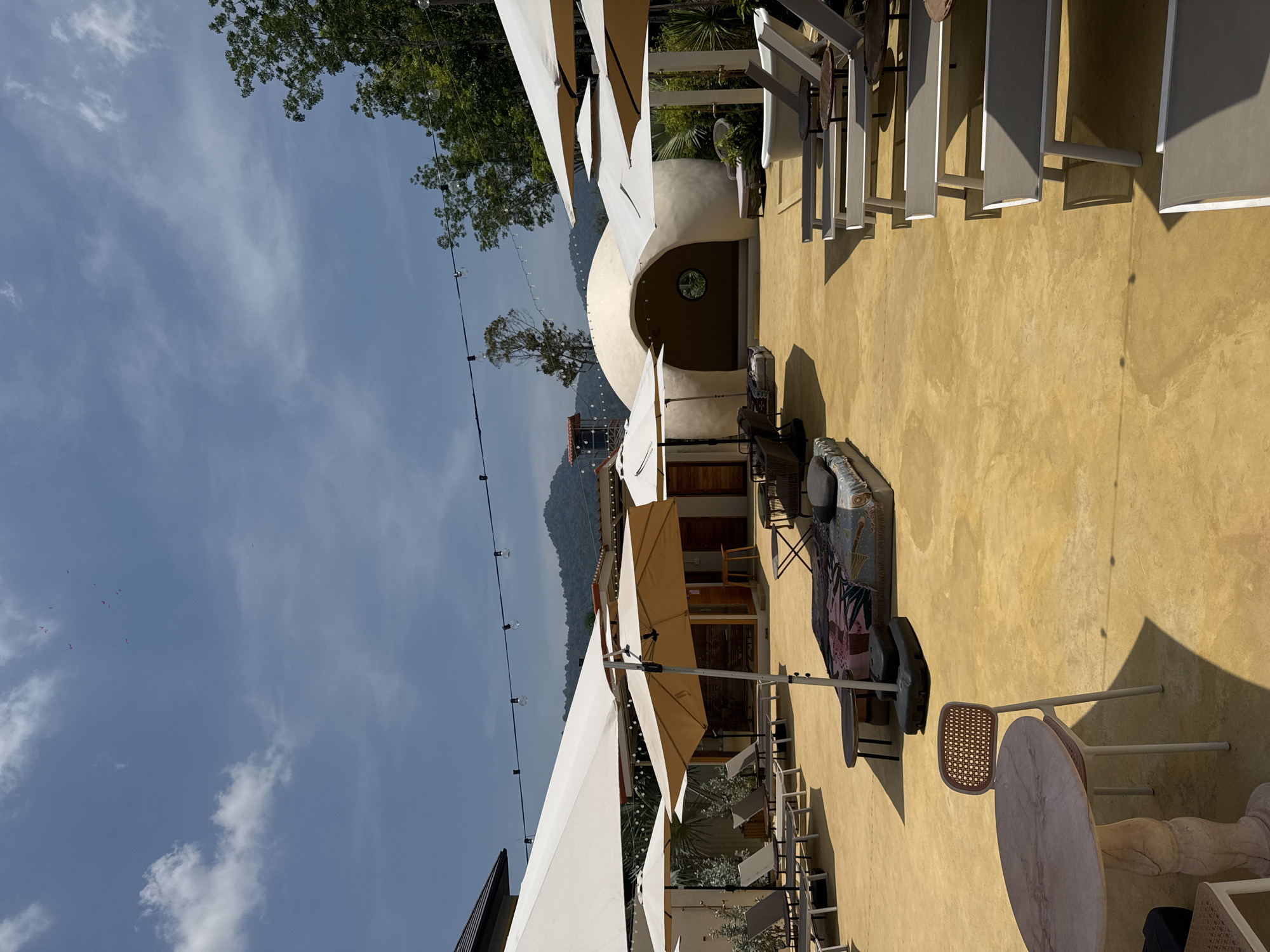 This is an outdoor space at the Baba Nest Rooftop Bar in Thep Krasatti, Thailand. The area is a patio with a golden surface. A low, dome-shaped structure with a round window is in the background. Various seating arrangements are present, including loungers under shade structures, tables with chairs, and a lounge area with a sofa and cushions. String lights are visible overhead. The sky is visible to the left, along with a mountain range.