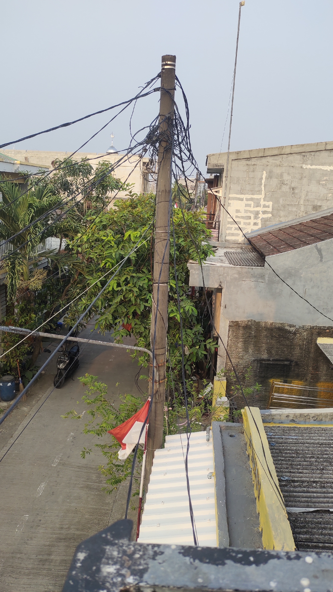 The image depicts an outdoor scene with a power pole prominently featured in the center. The pole is laden with a complex network of black electrical wires. To the left of the pole, a street is visible with a black motorcycle parked on it. Lush green trees with dense foliage surround the street and the base of the pole. A small red and white flag, likely the Indonesian flag, is attached to the pole, slightly to the left of its center.
In the background, several buildings are visible. One building on the left has a rounded, silver dome, possibly indicating a mosque. Other buildings appear to be residential or commercial structures with concrete walls and tiled roofs. The sky above is a pale blue, suggesting it might be early morning or late afternoon. The overall environment suggests a dense urban or suburban area in Indonesia, consistent with the location of Jalenjaya. The image is taken from an elevated perspective, looking down on the street and surrounding rooftops.