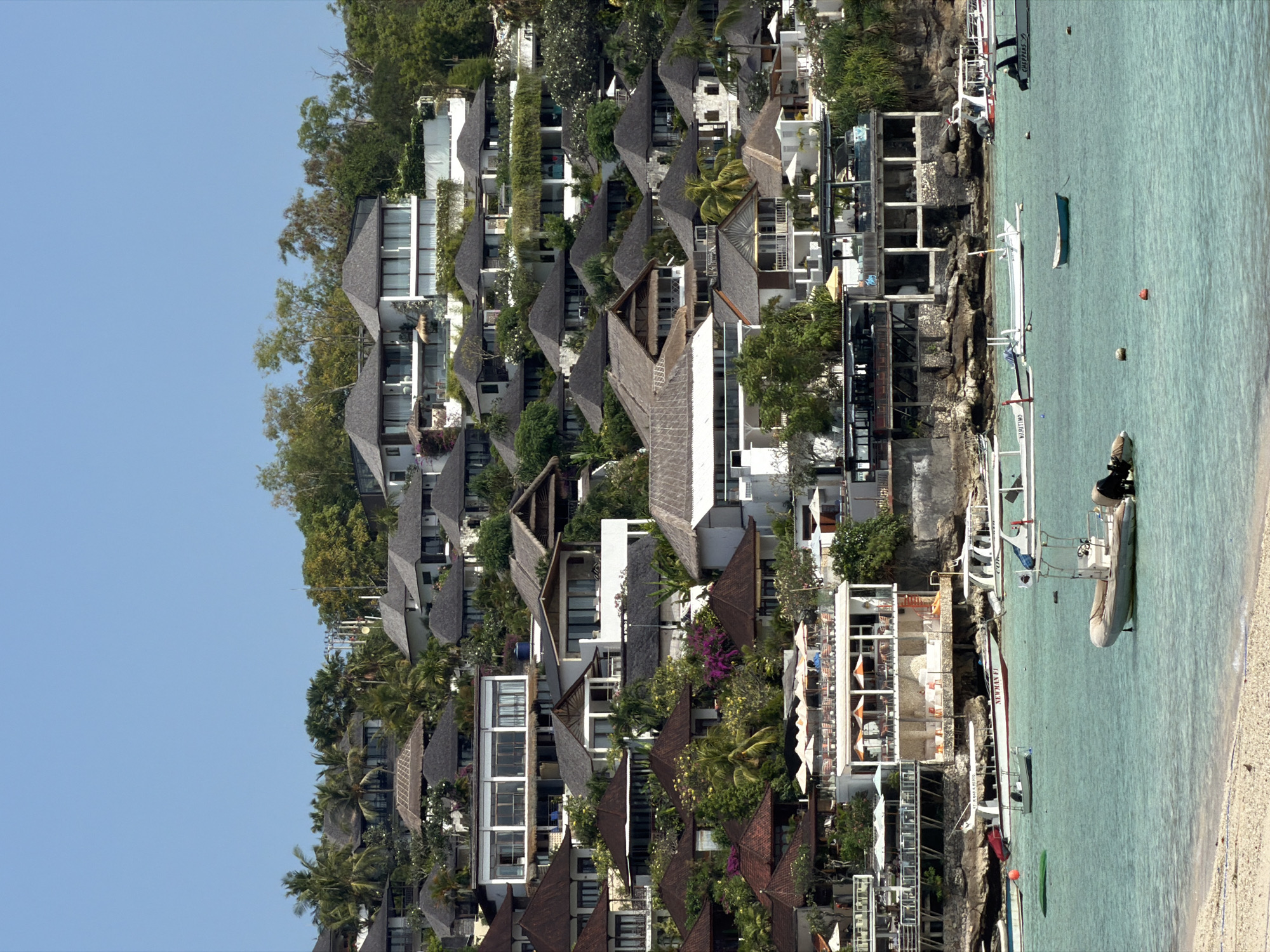 This is a medium-shot, vertical, daylight, eye-level, exterior photograph of a tropical hillside resort village on the coast of Nusa Penida, Indonesia. The scene depicts a dense collection of white buildings with dark grey, gabled roofs, nestled amongst lush green trees and palm trees on a steep incline. The resort overlooks a calm, turquoise body of water where several small boats are moored. The buildings feature open balconies, large windows, and thatched roof structures, suggesting a warm climate and an emphasis on outdoor living. In the foreground, a sandy beach is visible along the water's edge. The sky is clear and blue.