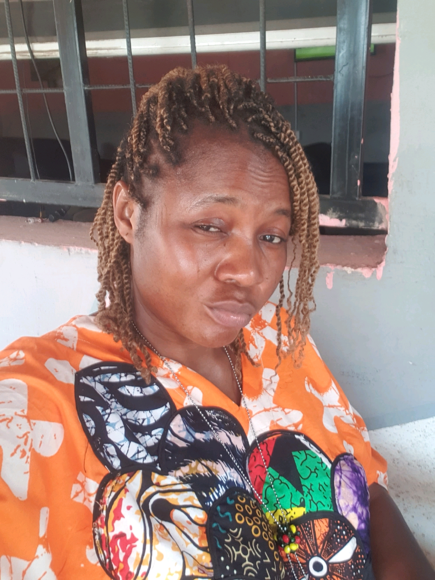 In Mangu Town, Nigeria, a woman with braided hair poses for a photo outdoors. She is wearing an orange shirt with a colorful, abstract pattern. She is looking towards the camera with one eye partially closed. In the background, there appears to be a building with bars on a window.