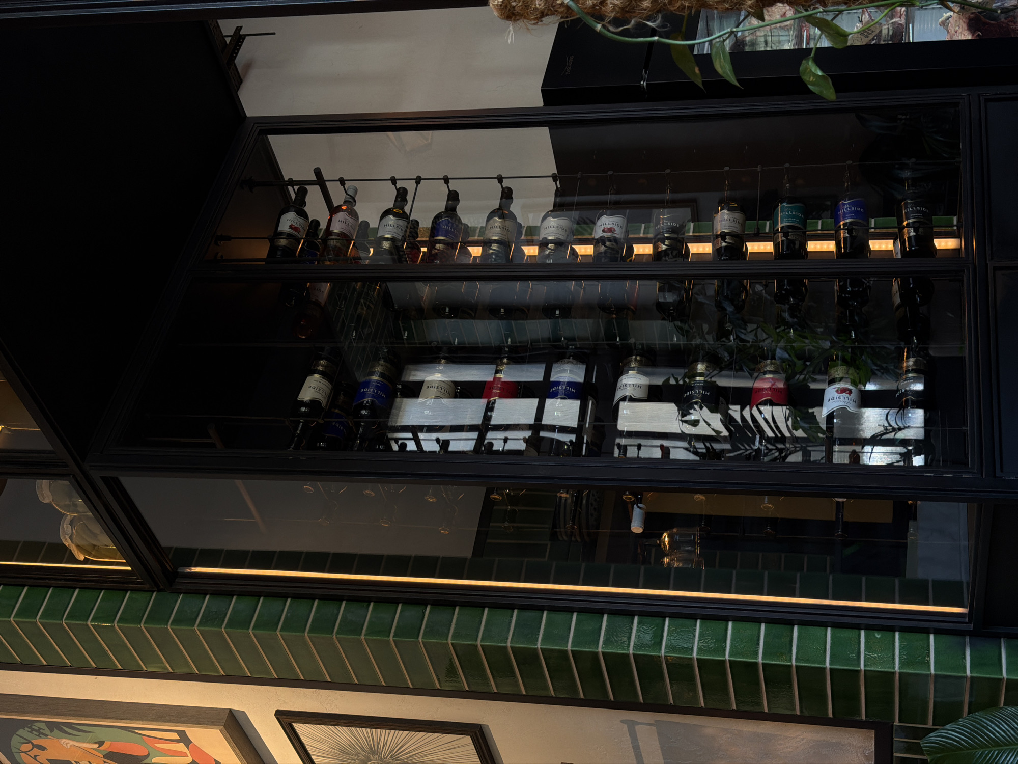 In a well-lit interior space, likely a restaurant or bar in Sabail Raion, Azerbaijan, a tall, multi-tiered wine rack occupies the central focus. The rack is encased in black metal framing and clear glass, allowing a view of its contents. The rack is filled with numerous wine bottles, many with white and blue labels reading "HILLSIDE," and some with red labels. Between the shelves, vertical strips of warm yellow light illuminate the bottles. To the left of the wine rack, a corner of green, glossy tiled wall is visible, with a sliver of a framed artwork above it. To the right, a black appliance, possibly a refrigerator, is partially visible, and lush green foliage drapes down, suggesting an indoor plant. The overall impression is one of a modern, sophisticated establishment.