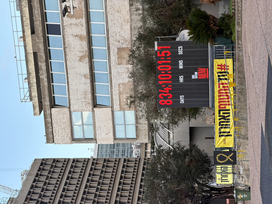 The image captures a public square in Tel Aviv, Israel, identified as Hostages Square (Kikar Hahatufim), located near the Tel Aviv District Court building. The central element is a large, black digital countdown clock, displaying "834:10:01:51" in prominent red LED numbers, beneath which are labeled "DAYS HRS MINS SECS". Below the numerical display, a red rectangular box contains the English text "BRING THEM HOME NOW".
In front of the digital clock, a long, yellow banner stretches across the lower mid-ground, featuring Hebrew text which translates to "#THELASTCHANCE" and is adorned with yellow fabric strips hanging from its lower edge. To the left of this banner, a smaller yellow sign displays a black background with a yellow ribbon motif.
On the left side of the frame, a tall, multi-story brutalist-style building, the Tel Aviv District Court, is visible with its characteristic repetitive grid-pattern facade. A construction crane is partially visible above its roofline. To the right of the digital clock, a multi-story building with a lighter-toned, textured brick facade and multiple rows of rectangular windows rises. A metal railing is present on its rooftop. The foreground consists of a paved plaza area. Several olive trees and other shrubs are interspersed throughout the scene. A green trash bin is visible on the far left. The sky is clear blue. No people are visible.