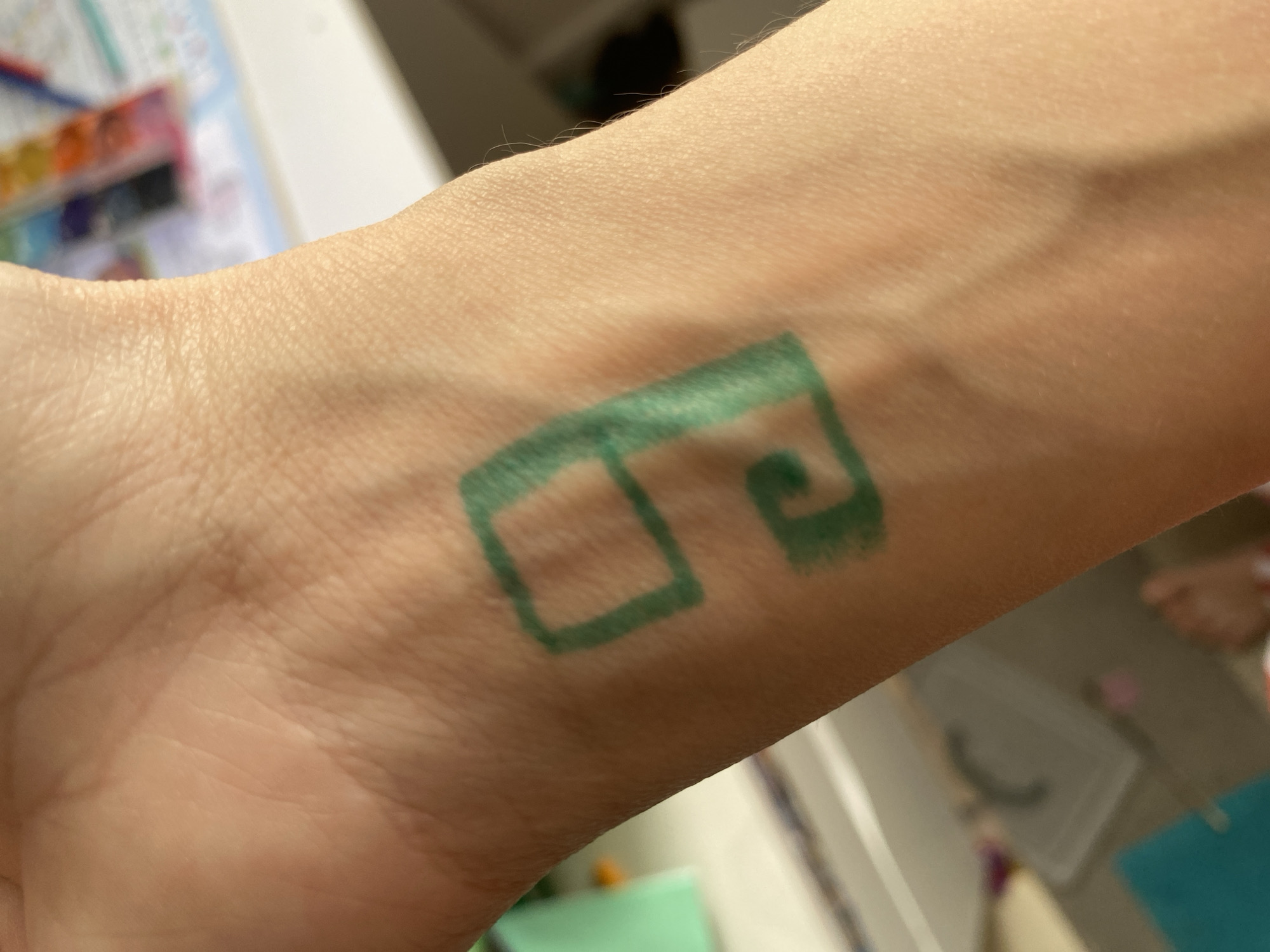 A human left forearm and wrist are depicted from a top-down perspective, displaying a green, stylized mark drawn on the volar surface. The mark resembles an open-ended number "9" or an abstract spiral, exhibiting pigment characteristics consistent with a temporary drawing implement. The skin shows typical epidermal texture and fine hair. The background, out of focus, indicates an indoor environment with a light-colored surface, likely a desk or table. Visible objects in the background include a watercolor paint palette containing multiple colors, a turquoise object, and a partially obscured human foot with a striped garment, resting on a light-colored floor next to a white container. The scene is evenly lit. Location: city of Unknown, Unknown.