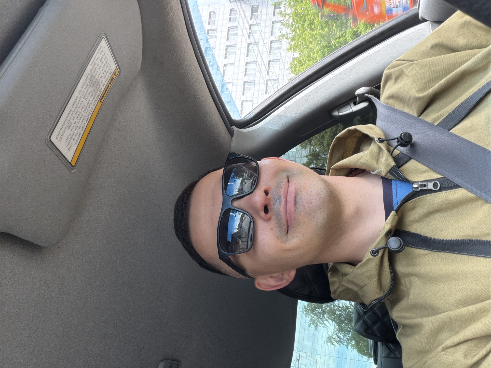 A man wearing sunglasses and a khaki jacket is inside a car. He is wearing a seatbelt. The car's sun visor is visible, as is a warning label on it. Through the car window, a building can be seen, suggesting an urban environment. Without additional information or context, it is not possible to confirm the location or city.
