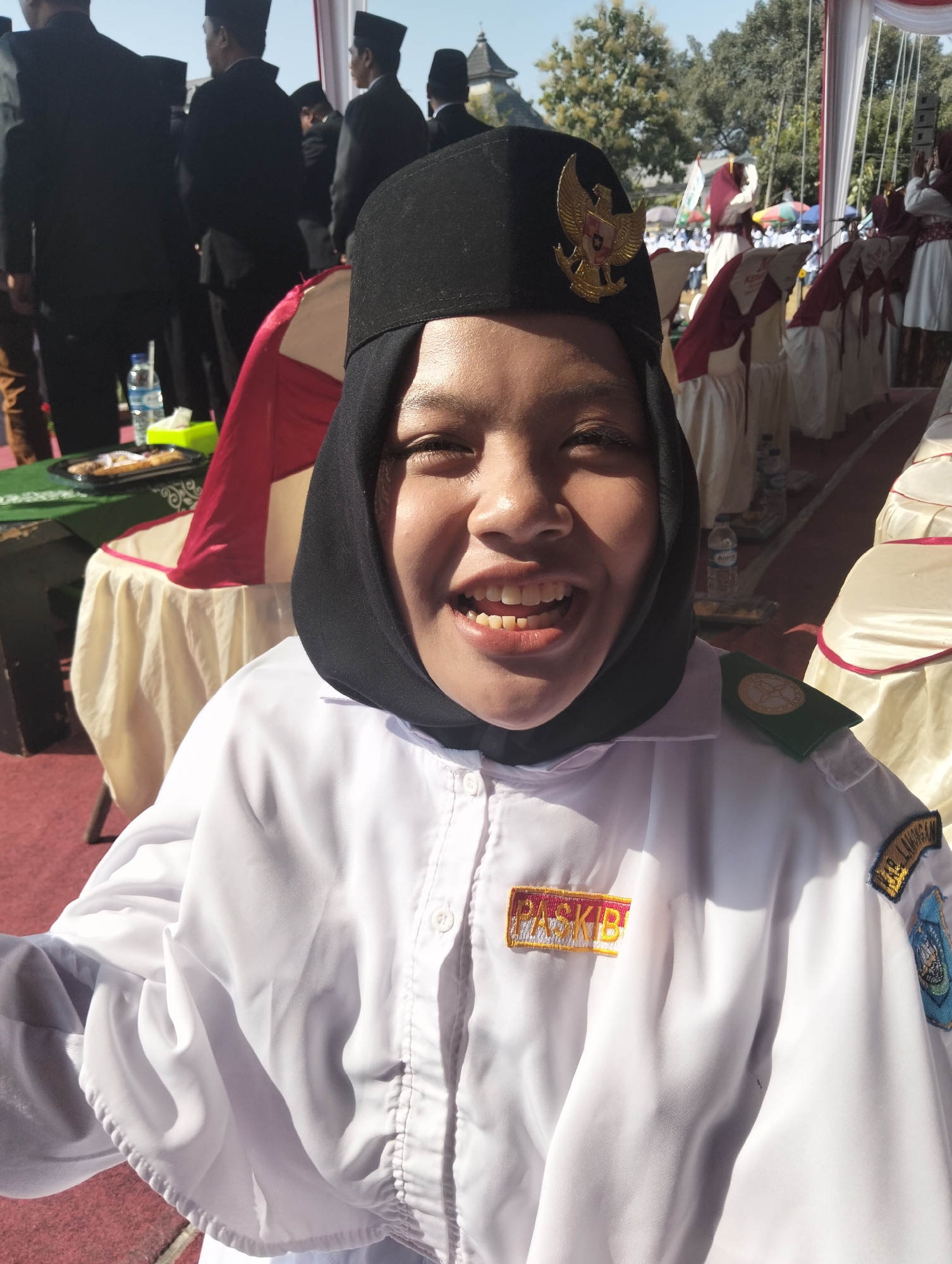 A young woman wearing a black hijab, black cap with a gold eagle emblem, and a white uniform shirt smiles broadly at the camera. The uniform shirt has patches on the shoulders and chest. She is outdoors in what appears to be a formal event. In the background, several people in black suits and caps are visible, along with white chairs covered with red and white fabric, and a red track. The scene suggests an outdoor ceremony or gathering in Paciran, Indonesia.
