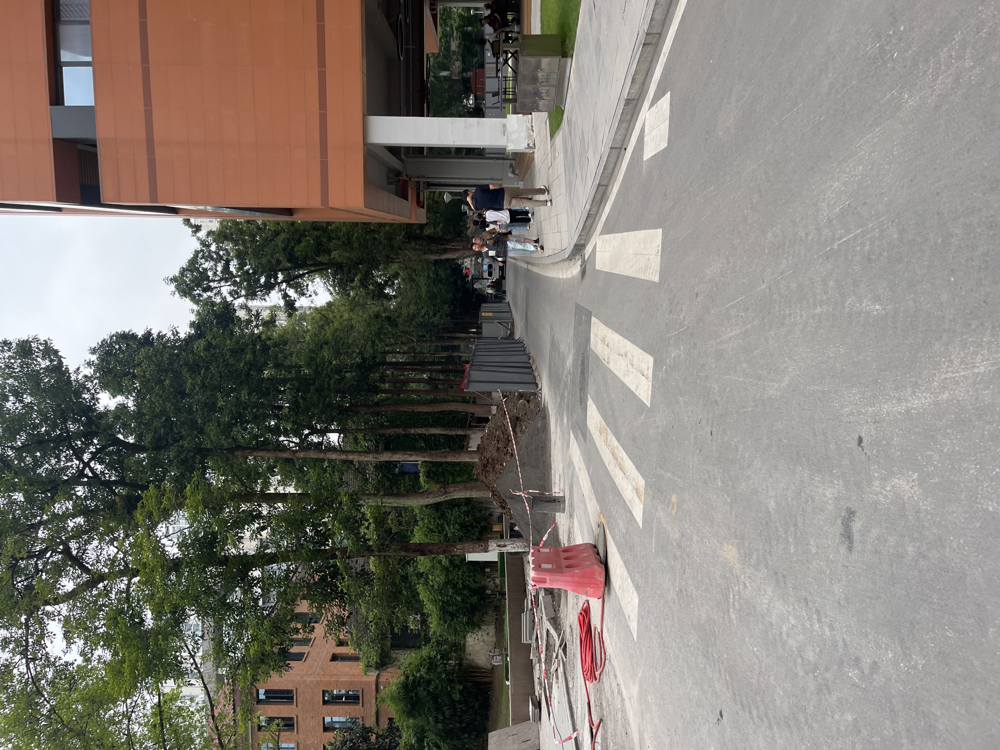 The image depicts a street scene in Yuzhong District, China, during daylight hours. The foreground features a road with a pedestrian crossing marked by white stripes. To the left of the crossing, construction materials are present, including a pile of dark sand, red construction barriers, and coiled red hoses. A concrete barrier with red and white tape is also visible.
Further down the road, several individuals are walking. On the right side of the road, a modern building with an orange facade and white pillars is prominent. Trees line the left side of the road, with a brick building partially visible behind them. The sky is overcast. The scene appears to be an outdoor public space undergoing some form of construction or maintenance.