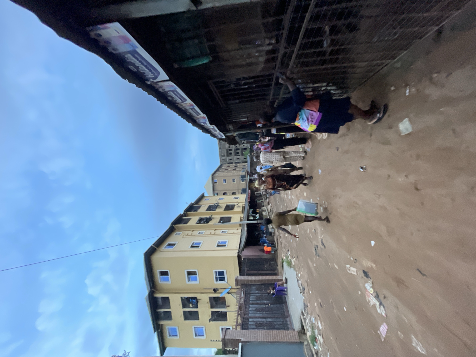 The image shows an outdoor scene in Nkpor, Nigeria. It depicts a street lined with buildings and vendors. A yellow, multi-story building is on the left. People are walking on the street, some carrying items. Vendors are seen along the edge of the street under a roofed, caged structure. The ground is brown and littered with debris. The sky is blue.