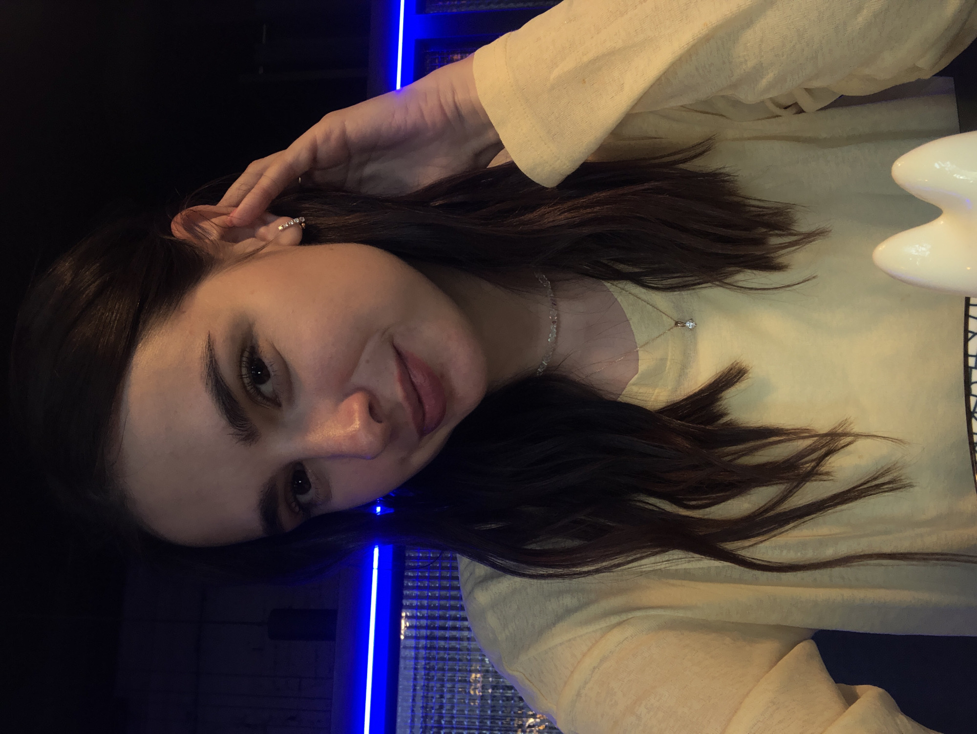 The image shows a young woman with long, dark hair, wearing a light yellow long-sleeved shirt and a delicate necklace with a small pendant. She is posing for a selfie, with her right hand touching her ear, and she is smiling slightly. The background appears to be a dimly lit interior, with a blue neon light strip visible. There is no identifiable information to suggest the location is in Izhevsk, Russia.