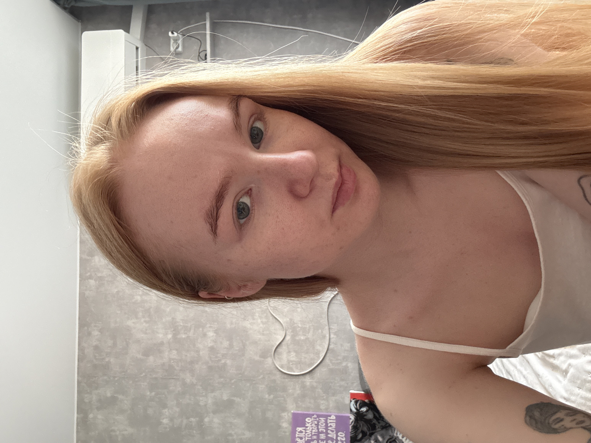 The media file presents a close-up portrait of a young woman with long, straight, light blonde hair and fair skin. The image is rotated 90 degrees clockwise, showing her head tilted to the left. She has light green or hazel eyes and a neutral expression, gazing directly towards the camera. She is wearing a white or off-white camisole with thin straps, and a tattoo is visible on her right upper arm.
The background indicates an indoor, domestic setting. A grey, textured wall serves as the primary backdrop, with a plain white wall section to the left. In the upper right background, white cables and a light-colored fixture are visible. A distinctive white cable shaped like a heart is arranged on the grey wall behind her shoulder. In the lower left, a purple object, likely a book or box, features partially visible text in Cyrillic script. Recognizable words include "ТОЛЬКО" (only), "ТВОРИ" (create), and parts of phrases like "В ЭТОЙ" (in this) and "ДЕЛАТЬ" (to do), confirming Russian text, which aligns with the provided context of Chaikovsky, Russia. The lighting is soft and even, suggesting it is daytime. No specific activity is depicted beyond posing for the camera.