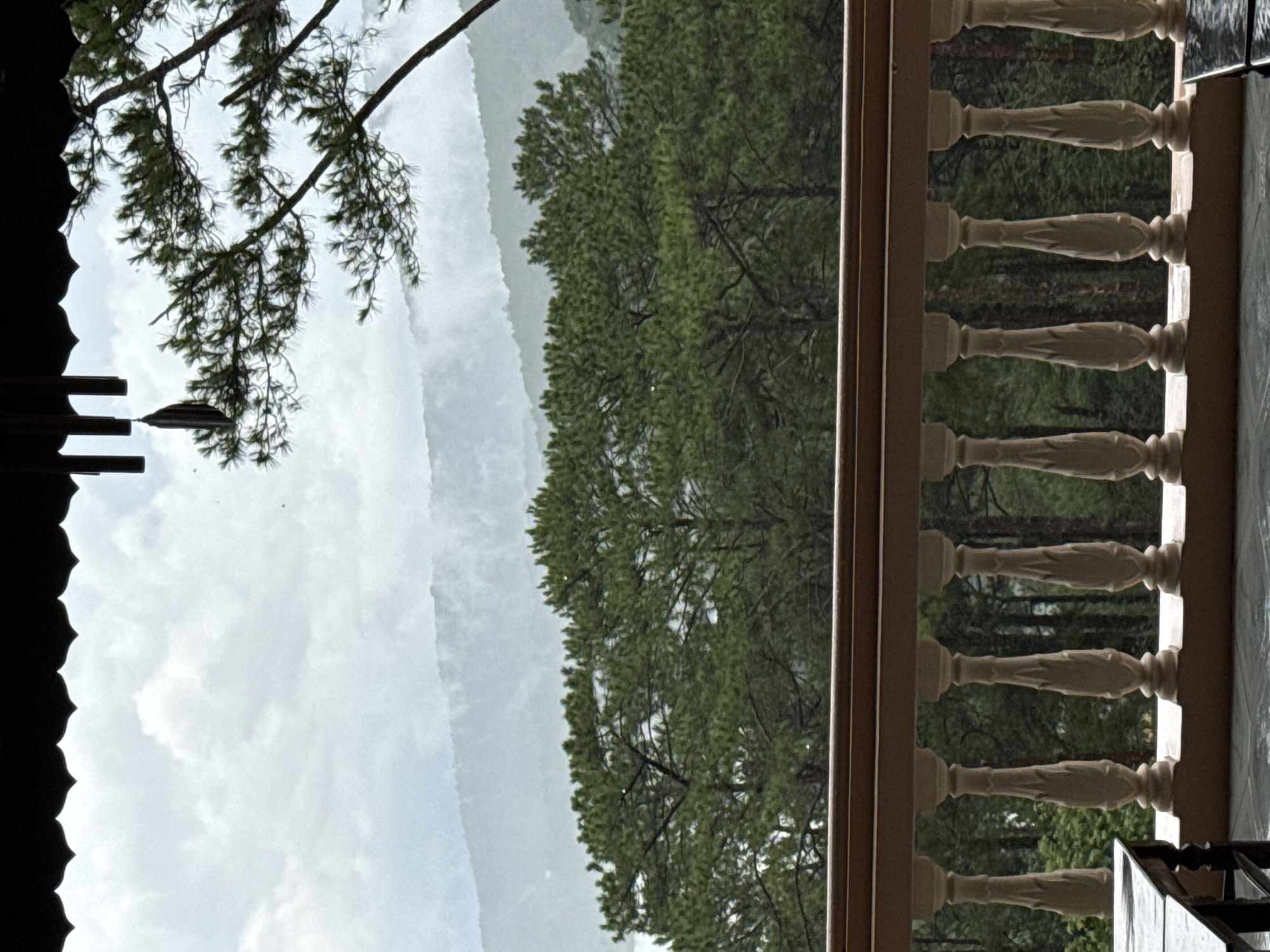 This image depicts a view from a balcony in Da Lat, Vietnam, overlooking a misty mountain landscape and a dense forest of pine trees. The foreground features a decorative beige wooden railing with intricately carved balusters. To the left, a portion of a dark wooden table with a shiny surface is visible. The middle ground is dominated by the lush green canopy of pine trees. In the background, rolling hills are shrouded in a soft, atmospheric mist, with layers of mountains fading into the distance under a cloudy sky. The roof of the structure from which the photo is taken, with a scalloped edge, is visible at the very top of the image, along with some dark hanging ornaments that resemble chimes. The overall scene conveys a sense of natural tranquility and a cool, humid climate, typical of Da Lat's mountainous terrain.