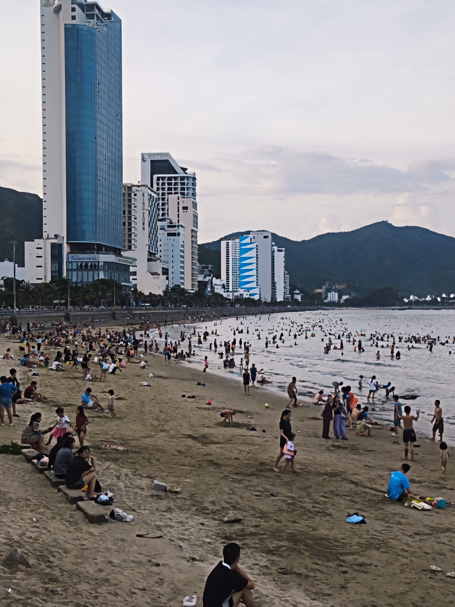 A wide-angle, eye-level shot captures a bustling beach scene in Nha Trang, Vietnam. The foreground shows a sandy beach with scattered debris and groups of people relaxing and playing. A few individuals are seated on low concrete blocks near the edge of the beach. Further back, a large number of people are wading and swimming in the ocean. The middle ground reveals a crowded shoreline with many people of various ages. In the background, a line of modern, tall buildings, including high-rise hotels and condominiums, stretches along the coast. The tallest building, a white and blue skyscraper, is prominently featured. Beyond the city buildings, a large, dark green mountain range forms the backdrop under a cloudy sky. The overall impression is one of a lively coastal city on a slightly overcast day.