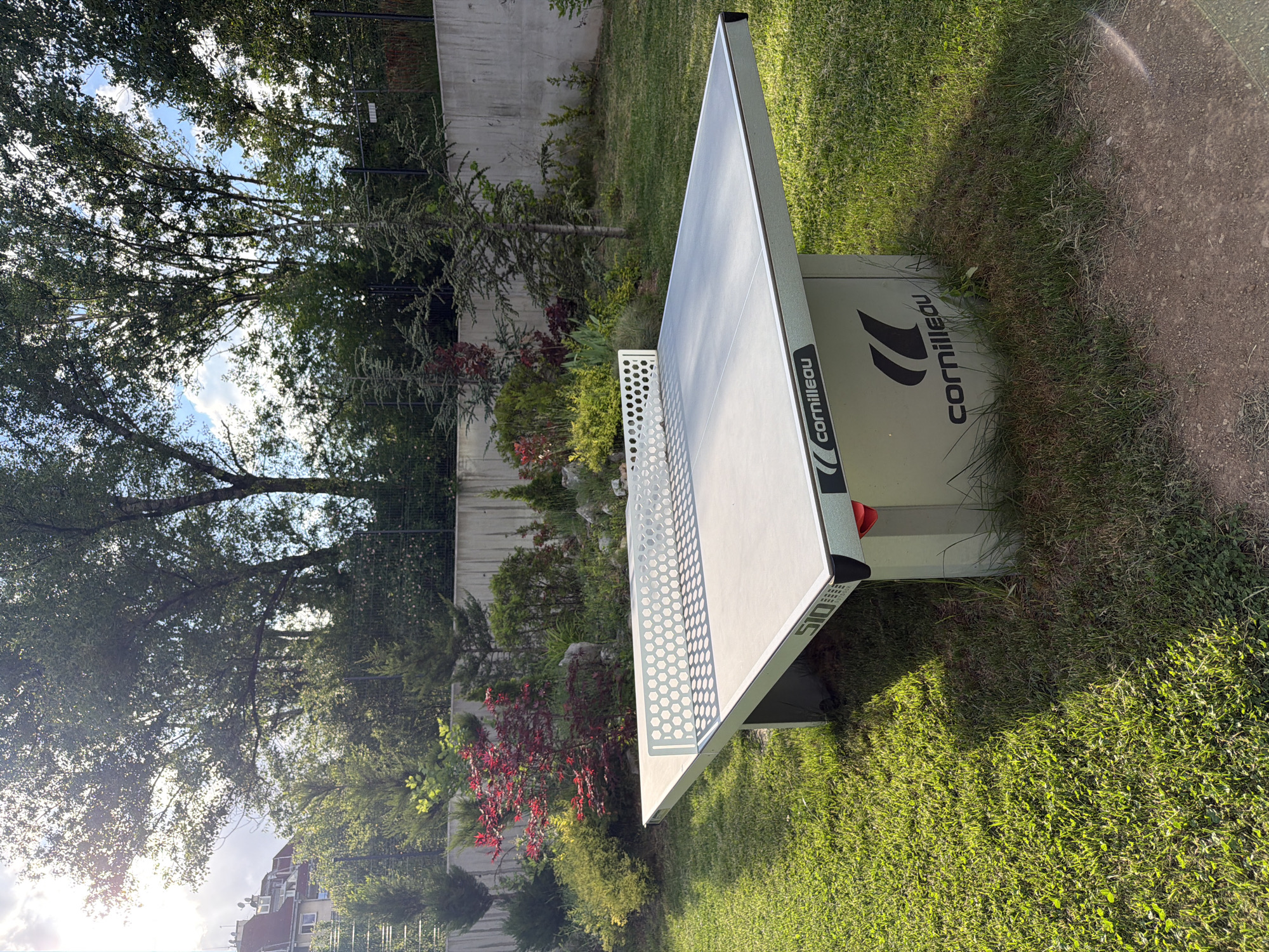 A Cornilleau ping pong table stands on grass outdoors. The table is light gray with black lettering and accents of red. The table sits in a garden setting with trees, bushes, red flowering plants, and a concrete wall in the background. A dirt path is to the right of the table. The photo was taken in Sofia, Bulgaria.