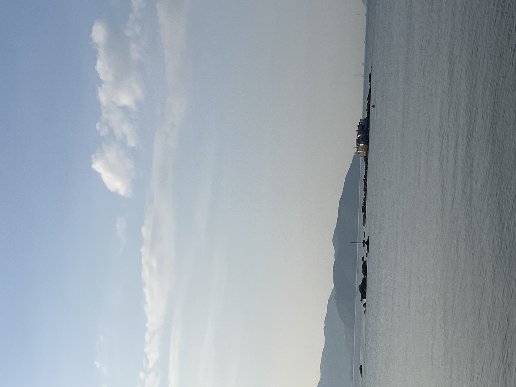 The image depicts a seascape, likely taken in or near Bac Nha Trang Ward, Vietnam. To the left, a blue sky is visible with streaks of white clouds. A mountain range lines the horizon, blending into the sky. To the right, a body of water occupies a significant portion of the frame. Some structures are visible along the shoreline, possibly buildings or piers. The overall scene suggests a coastal environment in Vietnam.