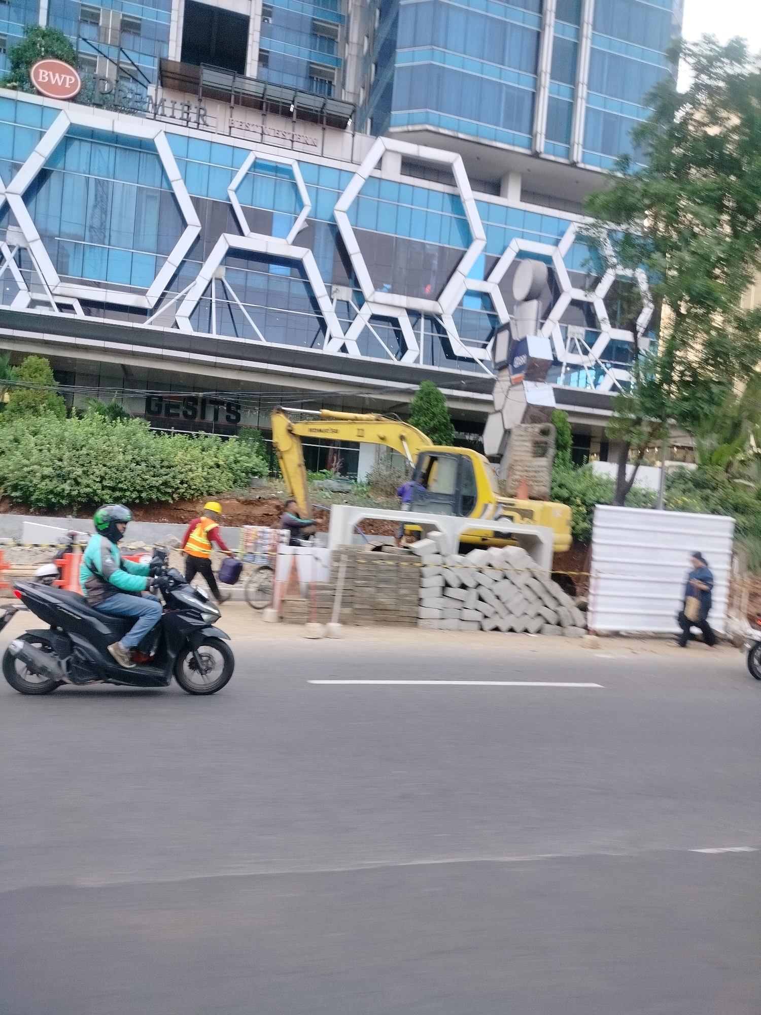 In East Jakarta, Indonesia, a motorcyclist wearing a teal jacket and a green helmet rides a black motorcycle on a paved road. Behind the motorcyclist, construction is underway. A yellow excavator is parked near a pile of concrete blocks. Two construction workers, one in an orange vest and yellow hard hat and another in a purple shirt, are present. A person in a dark jacket and light pants walks away from the construction area. A white corrugated metal barrier is installed on the side of the road. The background features a modern building with blue windows and hexagonal architectural elements, and lush green trees.