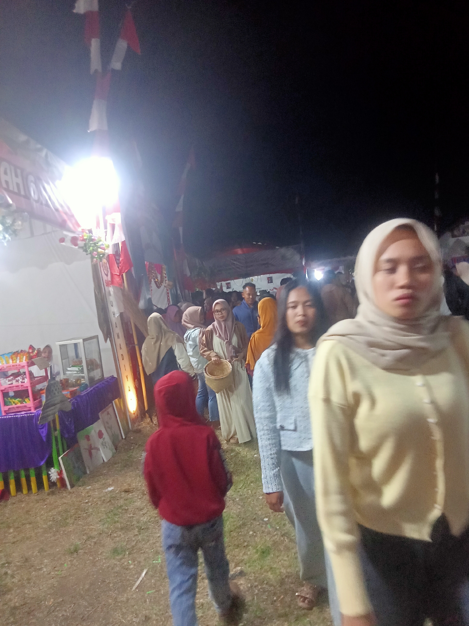 The image depicts an outdoor event at night, possibly a fair or market, in the city of Pule, Indonesia. Several people are visible, some in the foreground and others in the background. A young person in a red hoodie and blue jeans walks away from the camera. To their right, a woman wearing a light yellow hijab and sweater is in the foreground, partially obscuring another woman in a light blue jacket and gray pants. Other individuals, some in hijabs and traditional attire, are visible further back, milling about. Several stalls with colorful displays are set up, including one with a rack of items and another with artwork. Red and white flags, possibly national flags, are strung overhead, and a bright light source illuminates part of the scene. The ground appears to be covered in grass and dirt, indicative of an outdoor venue.