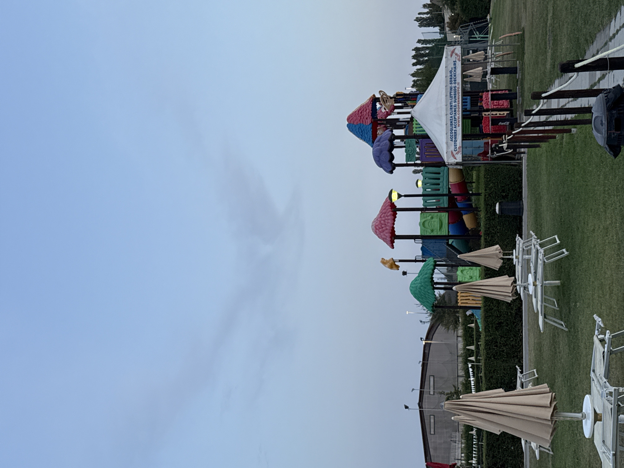 The image depicts a playground and a beach-like area at dusk in Parma, Italy. In the foreground, several folded beach umbrellas and lounge chairs are arranged on a grassy lawn. A paved pathway leads towards the right side of the image, marked by a wooden railing with rope accents. Further back, a colorful playground structure with slides and climbing elements stands prominently. A white tent with text in Italian and English is situated in front of the playground, advertising services such as "SUNBEDS - DECKCHAIRS". A large, gray building with visible lights is located in the background on the left, suggesting it might be part of a facility or complex. The sky is a pale blue, transitioning to a darker hue, indicating the late hour. There are no people visible in the scene.