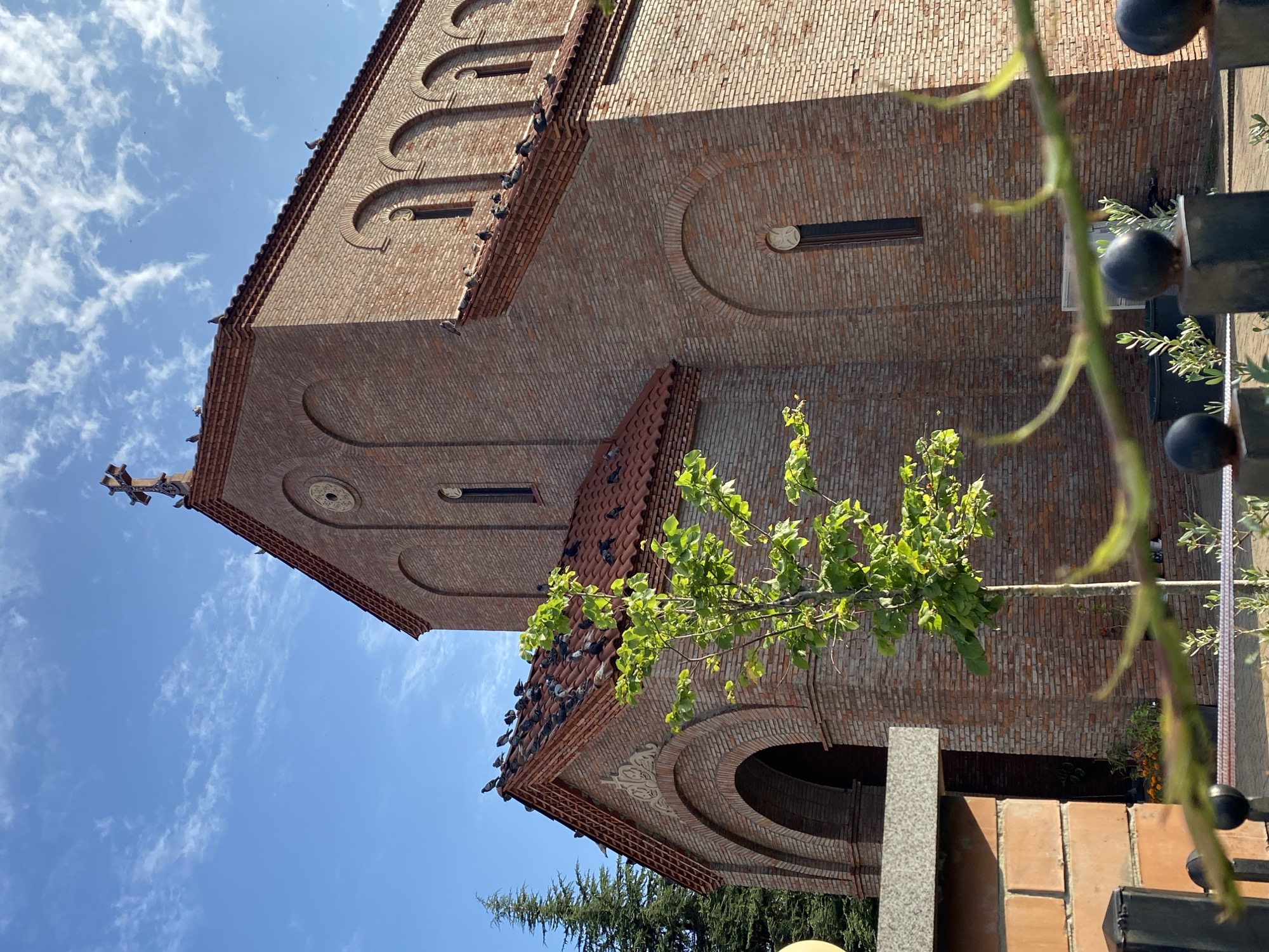 A brick church with a red tiled roof stands against a bright blue sky with scattered clouds. A cross adorns the peak of the roof, and several pigeons are perched on the roof's edge and near the cross. A small, leafy tree is positioned in front of the church, with its branches reaching towards the building. In the foreground, a black metal fence with round finials partially obstructs the view, and a brick wall is visible to the left. The scene appears to be outdoors, possibly in a courtyard or garden area. This church is located in Batumi, Georgia.