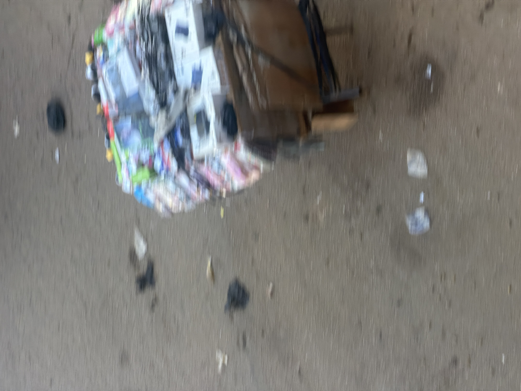A cart piled high with assorted goods, possibly for sale or being transported, is situated on a paved surface in Bukuru, Nigeria. The cart's wooden frame is visible, with dark metal legs and supports. Among the items are numerous white boxes, some with blue lettering, and a variety of colorful packaging. Several dark, circular objects are scattered on the ground near the cart, as well as some smaller debris. The ground appears to be asphalt or a similar paving material. The overall image has a blurred quality, suggesting motion or a lack of sharp focus.