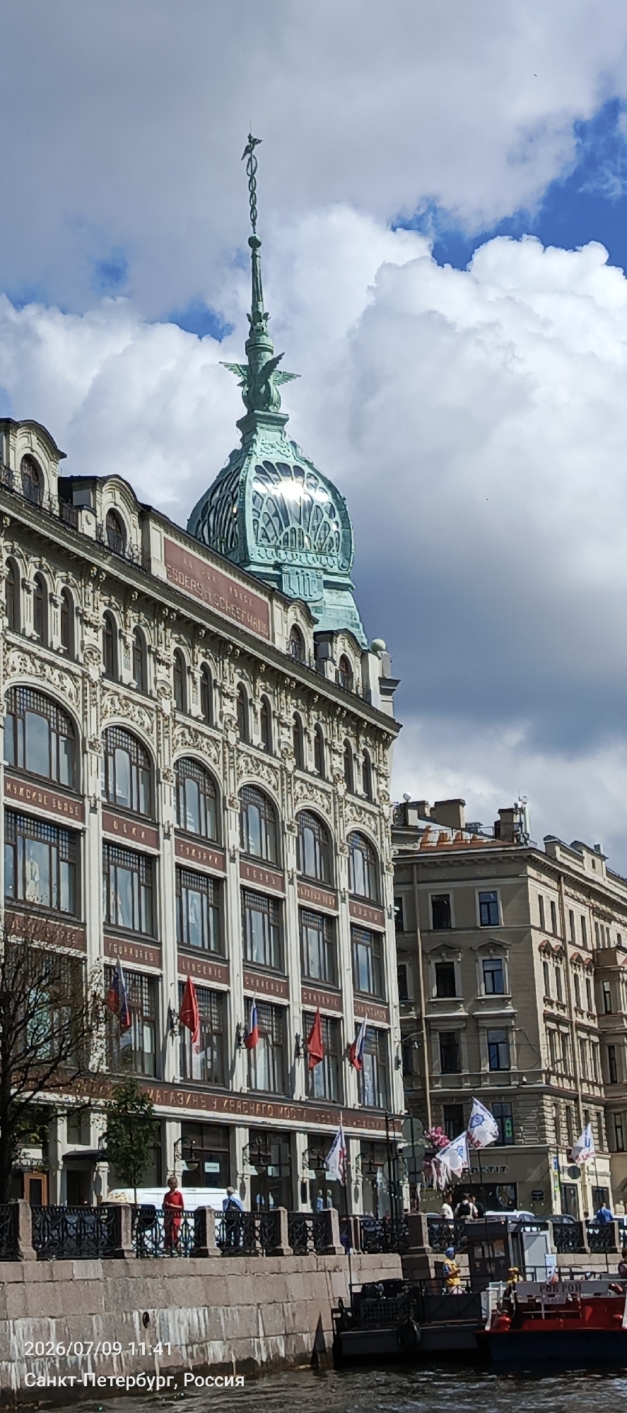The image shows a close-up view of the Singer House building in Saint Petersburg, Russia, on a cloudy day. The ornate facade of the building, with its numerous windows and decorative elements, is visible, as is the distinctive green dome topped with a statue of an eagle. Several flags are displayed along the building's facade, and a row of trees lines the embankment in front of it. Below the embankment, a body of water is present, with a boat docked and another partially visible. People are seen walking on the embankment and on the boat. The text at the bottom of the image indicates the date and location: "2026/07/09 11:41" and "Санкт-Петербург, Россия" (Saint Petersburg, Russia).