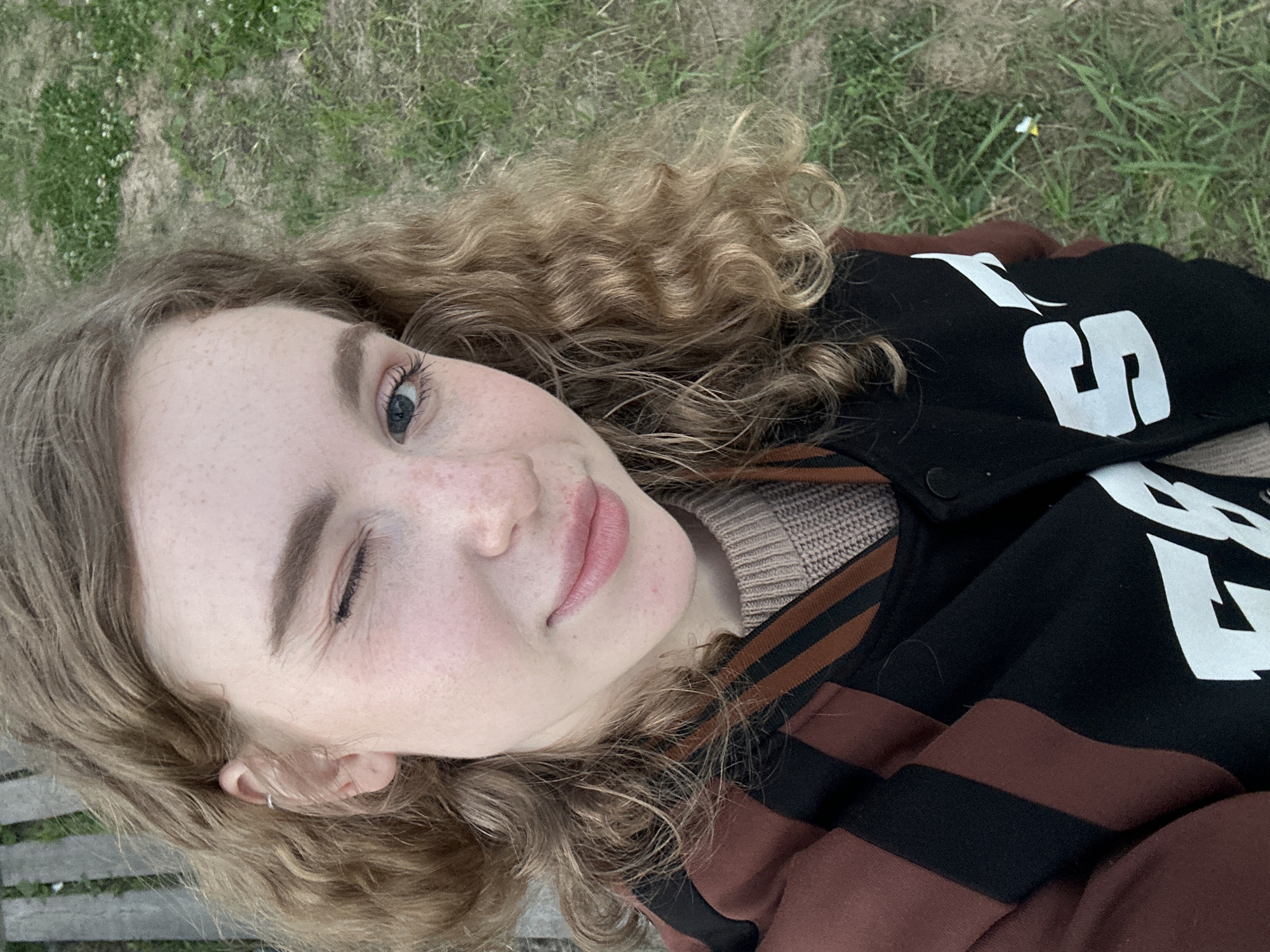 A close-up, high-angle selfie depicts a young person, likely female, lying outdoors on green grass in Vasilyevo, Russia. The subject has light, curly, shoulder-length blonde or light brown hair, fair skin with noticeable freckles, and blue eyes. They are winking with their left eye and displaying a subtle, closed-mouth smile, conveying a playful and relaxed expression. A small silver hoop earring is visible in their left ear.
The person is dressed in a black jacket featuring broad brown and black striped sleeves. Underneath, a light brown or tan ribbed knit sweater is visible. Large white text or numbers adorn the black body of the jacket; parts of an 'S' and a stylized '5' or '2' are clearly legible.
The background is dominated by green grass with some drier patches and a few small white or yellow flowers. A wooden surface, possibly a bench or part of a boardwalk, is partially visible behind the subject's head on the left side of the frame. The natural light suggests it is daytime, creating a bright and clear image.