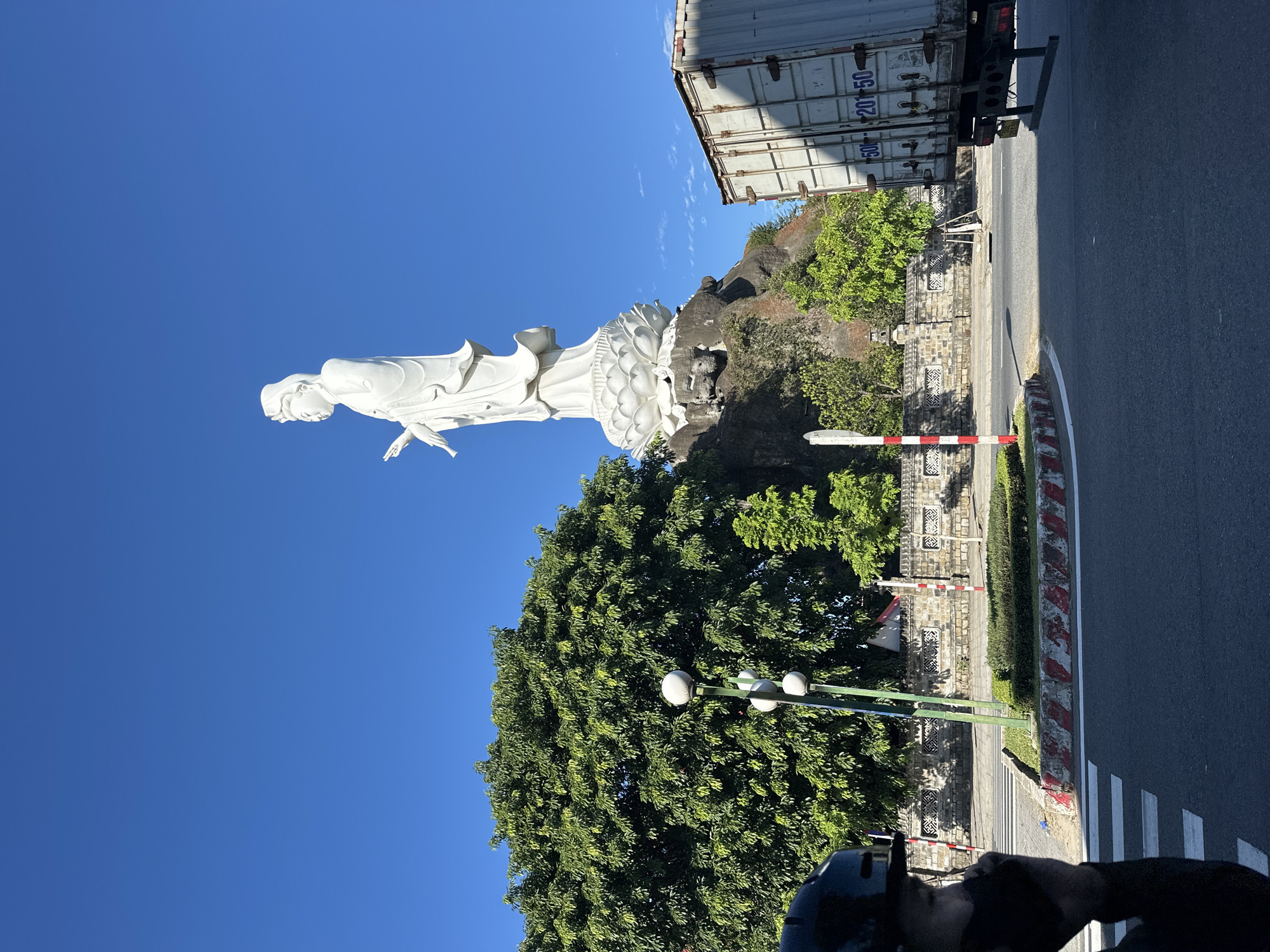 The image shows the Lady Buddha statue (Chùa Linh Ứng) in Đà Nẵng, Vietnam, a prominent landmark situated on Sơn Trà Peninsula overlooking the South China Sea. The white statue, depicting the Goddess of Mercy, stands tall on a lotus-shaped pedestal atop a rocky hill, bathed in bright sunlight under a clear blue sky. A large, lush green tree is positioned to the left of the statue, with smaller trees and vegetation in the foreground.
In the lower portion of the image, a street scene unfolds. A person wearing a helmet and mask is partially visible on the left, holding a phone to their ear, suggesting they are using it. Across the street, a large white shipping container truck is parked, its side displaying numbers and lettering. A pedestrian crossing is visible on the asphalt road, with striped markings and a concrete curb. A stone wall with intricate patterns runs along the background, with decorative pillars and fencing. Traffic lights and street lamps are also present. The overall environment appears to be a sunny day in a well-developed urban or tourist area.