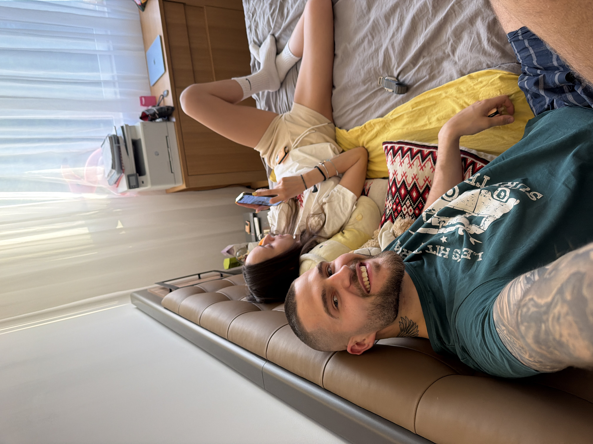 A man and woman are lying on a bed, oriented perpendicularly to each other in what appears to be an interior setting. The man, positioned in the foreground, is lying on his side, smiling toward the camera. He has short hair and a beard, and is wearing a dark green T-shirt and blue plaid shorts. His left arm, visible to the camera, has tattoos covering much of its surface. The woman, behind him, is lying on her back with her legs up in the air, feet in white socks, holding a mobile phone. She is wearing light-colored shorts and a shirt. They are lying on a bed with a brown leather headboard. The bed is covered in grey sheets, a yellow blanket, and a patterned pillow. A watch is visible on the sheets near her legs. In the background, a printer and a laptop are on a wooden dresser next to a bright window with sheer curtains. The room appears to be well-lit. There is no information available in the image to conclude it's taken in the city of Sofia, Bulgaria.