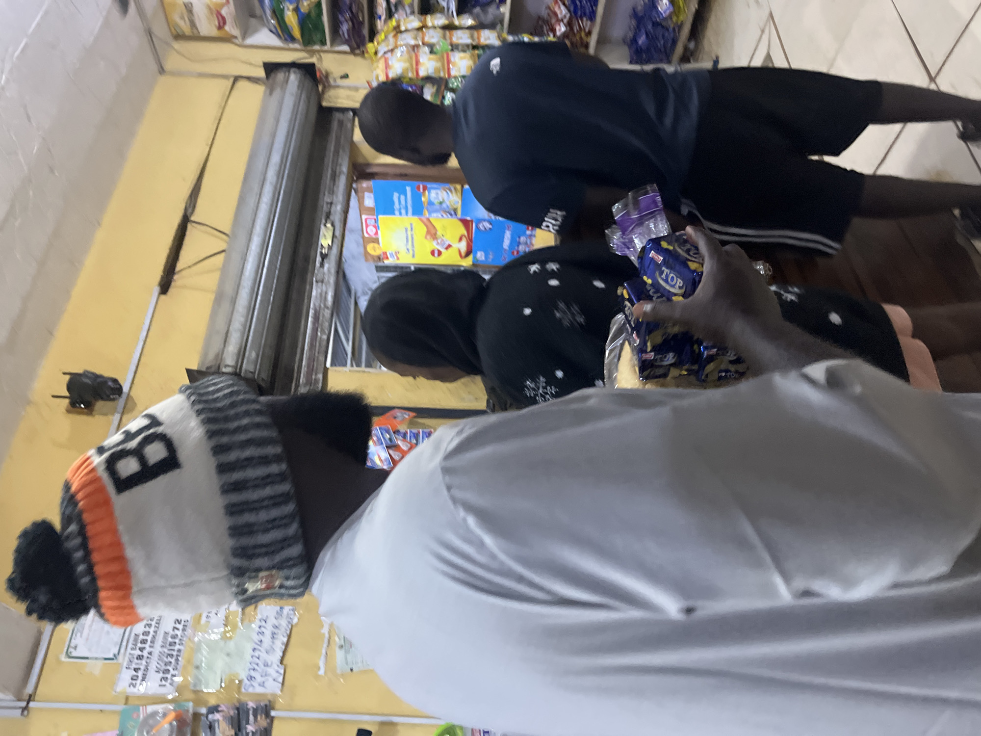 The image shows the interior of a small shop, possibly a convenience store, in Bukuru, Nigeria. Three individuals are visible. In the foreground, from the left, a person wearing a white long-sleeved shirt and a knit beanie with orange and black stripes is holding several packages of what appears to be biscuits or snacks. The beanie has the letter "BE" in black. Behind this person, a person in a black hooded garment is partially visible. To the right, a person in a dark blue t-shirt and black shorts stands facing away from the camera, looking into the shop. The shop interior is characterized by yellow walls and shelves stocked with various packaged goods, including what look like bags of crisps and other food items. A metal roll-up security gate is visible above the counter area. There are posters and signage on the walls, some with what appear to be bank details and advertisements. The floor is tiled. The overall impression is of a busy local shop.