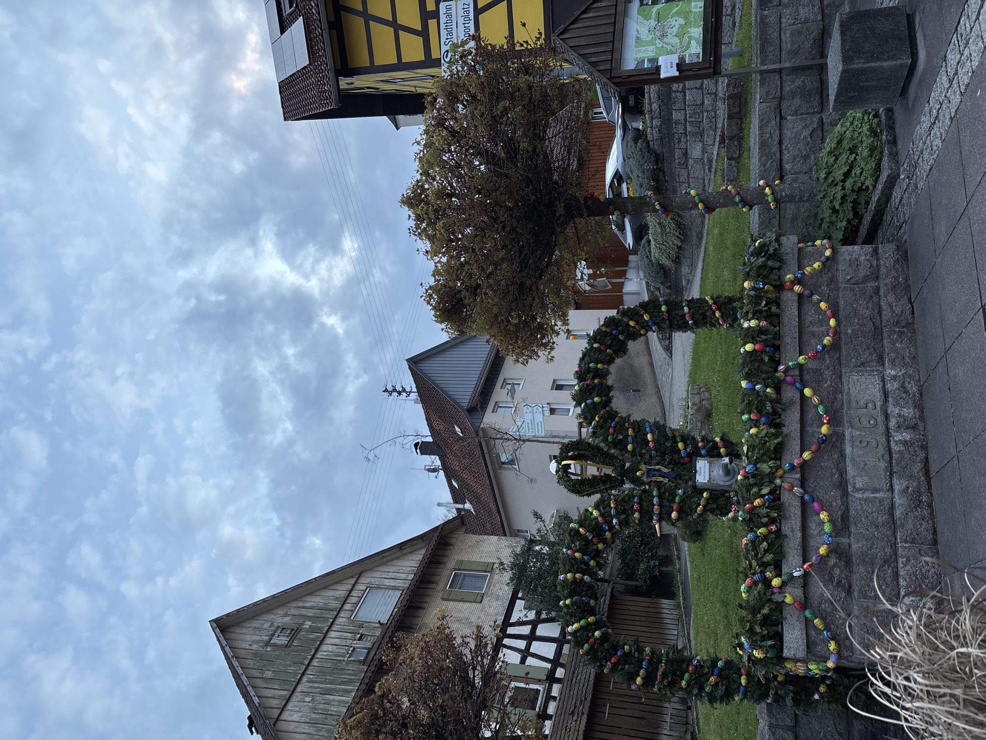 The image depicts a street scene in Forbach, Germany. The foreground showcases a stone structure adorned with garlands of greenery and colorful Easter egg decorations. Etched into the stone is the year "1965." A tree decorated with Easter eggs stands behind the structure. Behind the tree and stone structure is a grassy area.
The backdrop includes traditional German houses with pitched roofs and timber framing. An information board and street sign reading "Stadtbahn Sportplatz" are also visible. Above the houses, the sky is filled with gray clouds. Electrical lines are stretched across the sky in the upper left of the image. The image is taken from a ground-level perspective.