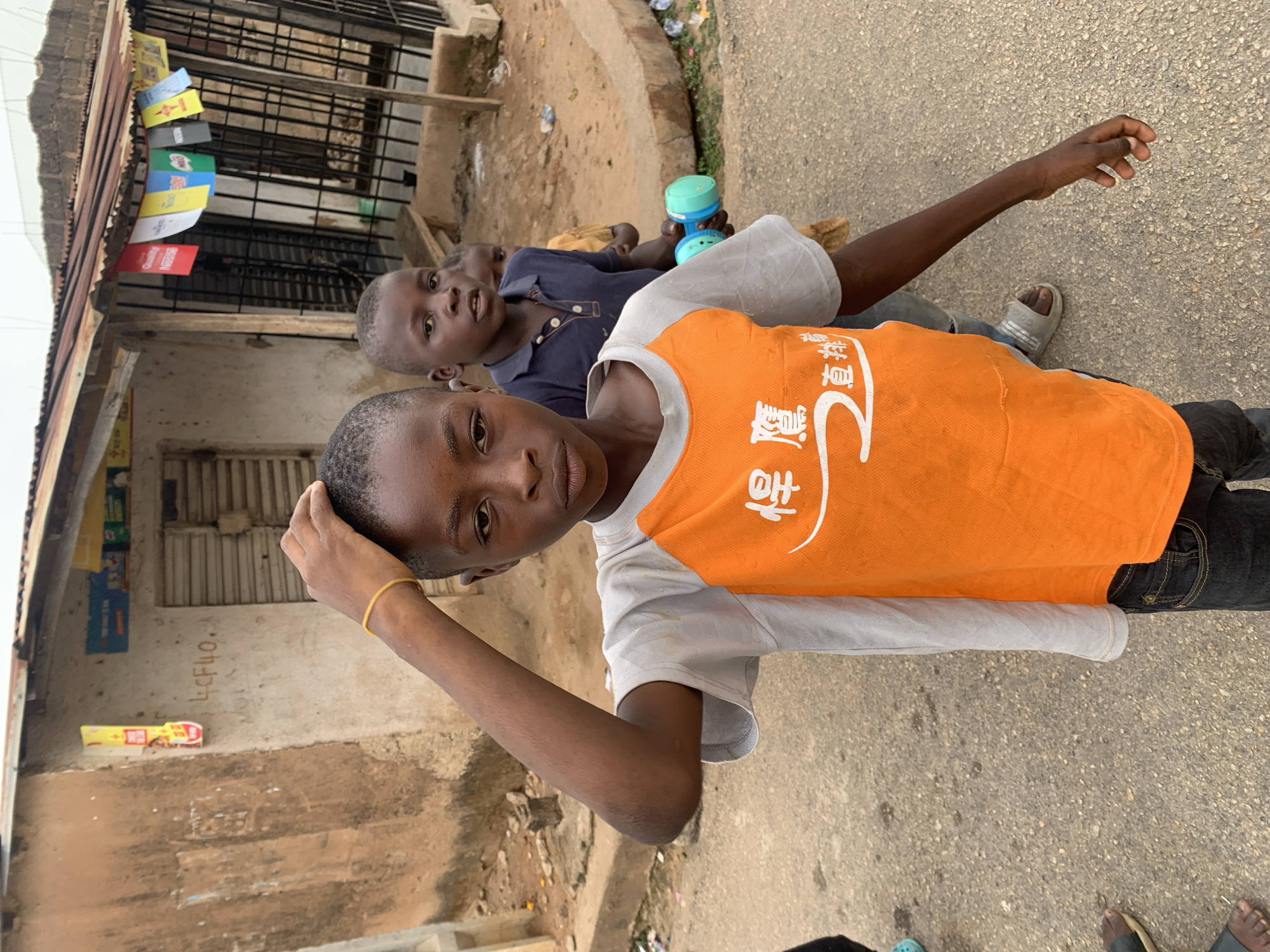 The image shows two young boys in an urban environment, likely a street in Jos, Nigeria. The boy in the foreground wears a grey and orange t-shirt with Chinese writing on it and has a gold-colored bracelet on his left wrist. His right hand is touching his head. He wears dark pants and sandals. The boy behind him wears a navy blue collared shirt and sandals, and is holding a blue and green object, possibly a toy.
They are standing on a paved surface that appears to be a road or sidewalk. Behind them is a building with a rusted corrugated metal roof. The building has metal bars over the windows and various signs and graffiti on the wall. There are various commercial promotional flags are hanging above the entrance. There is also trash and debris scattered on the ground.