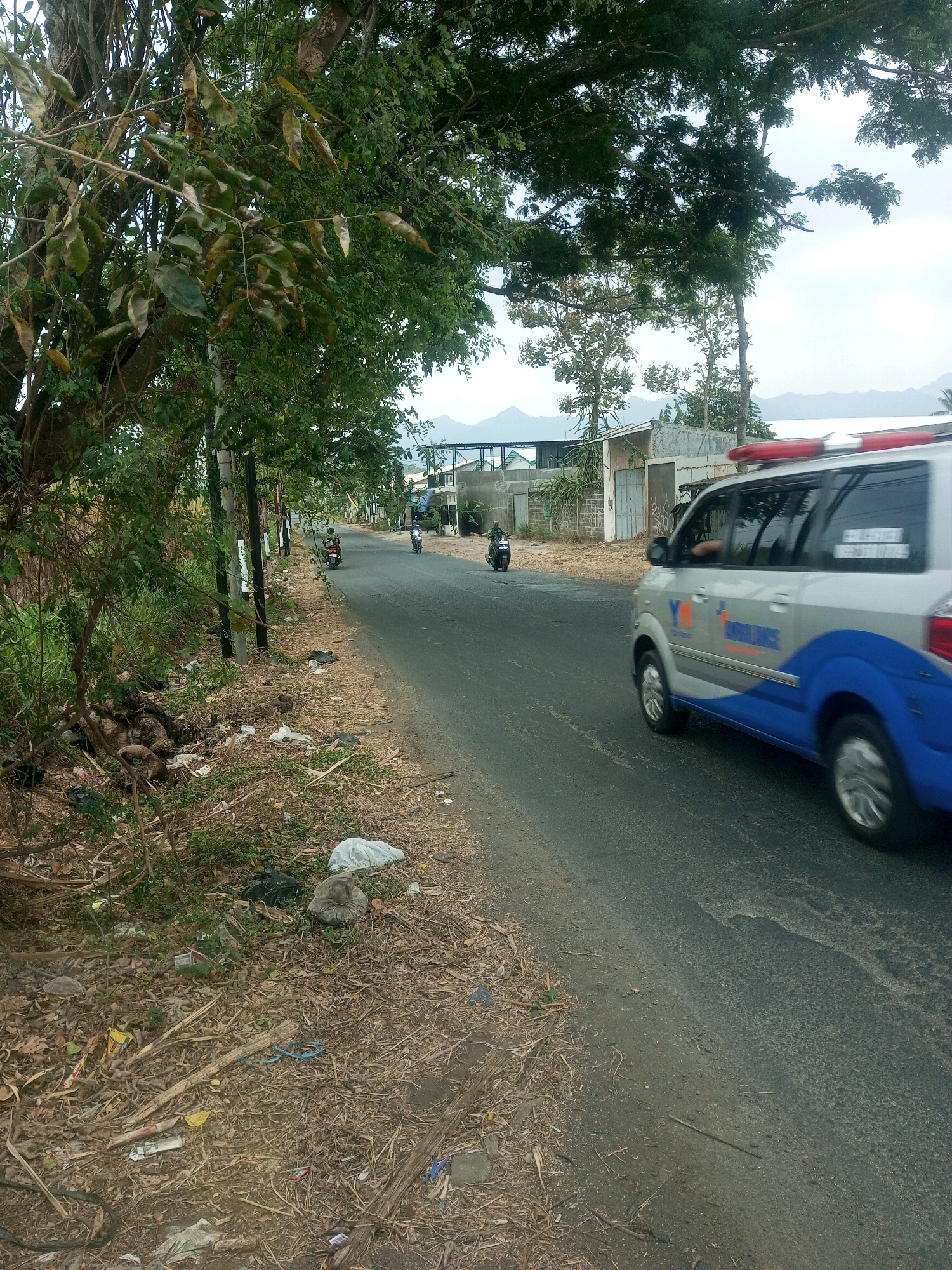 In Pakis, Indonesia, an ambulance van with blue and silver markings is driving on an asphalt road. The van has red flashing lights on its roof. A person is visible inside the van. To the left of the road, there is a grassy area with scattered debris, including plastic bags, dry leaves, and branches. Further down the road, two motorcycles are visible, one with a rider wearing a helmet. The background shows a few buildings with corrugated metal roofs and some trees. In the distance, mountains can be seen under a cloudy sky. The scene appears to be a typical roadside environment in a semi-urban or rural area.
