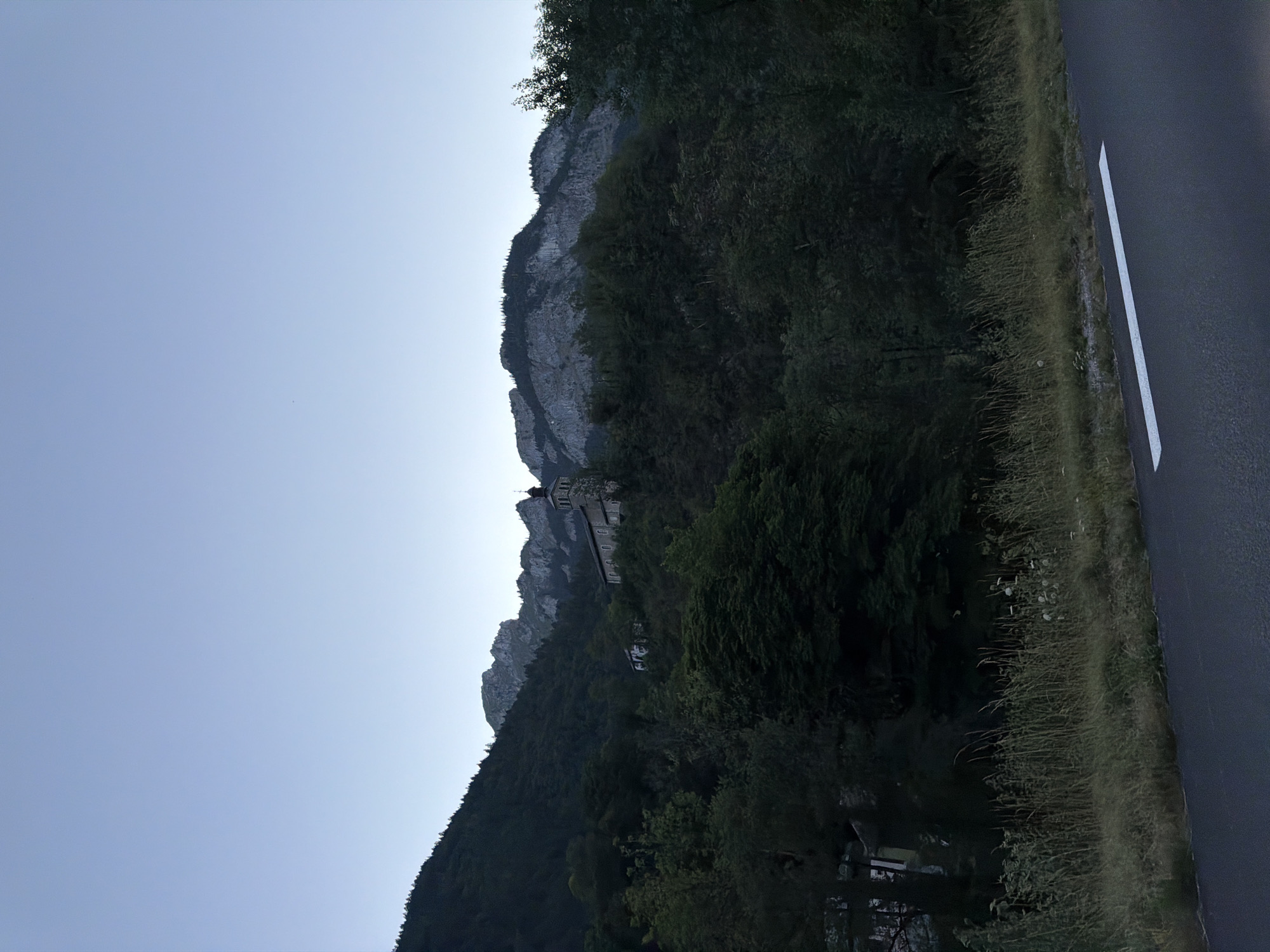 The image depicts a serene, twilight scene of a mountain landscape in La Baume, France. In the foreground, a grey asphalt road with a white line runs horizontally across the bottom of the frame, bordered by tall, dry grass and some wildflowers. The middle ground is dominated by a densely wooded hillside covered in various shades of green foliage, with a few scattered buildings visible amongst the trees. Perched higher on the hillside is a stone church with a tall steeple and a bell tower. Behind the church, imposing grey mountains with rugged peaks rise sharply against a pale blue, cloudless sky, indicative of early morning or late evening. The overall impression is one of natural beauty and tranquility.